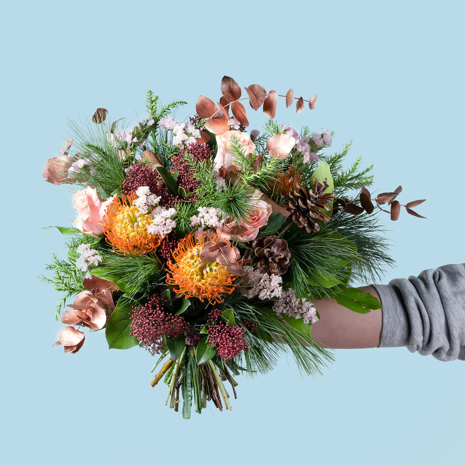 Ein farbenfroher Strauß verschiedener Blumen, darunter Rosen, Chrysanthemen und Tannenzweige, wird vor einem schlichten blauen Hintergrund in den Händen einer Person gehalten.