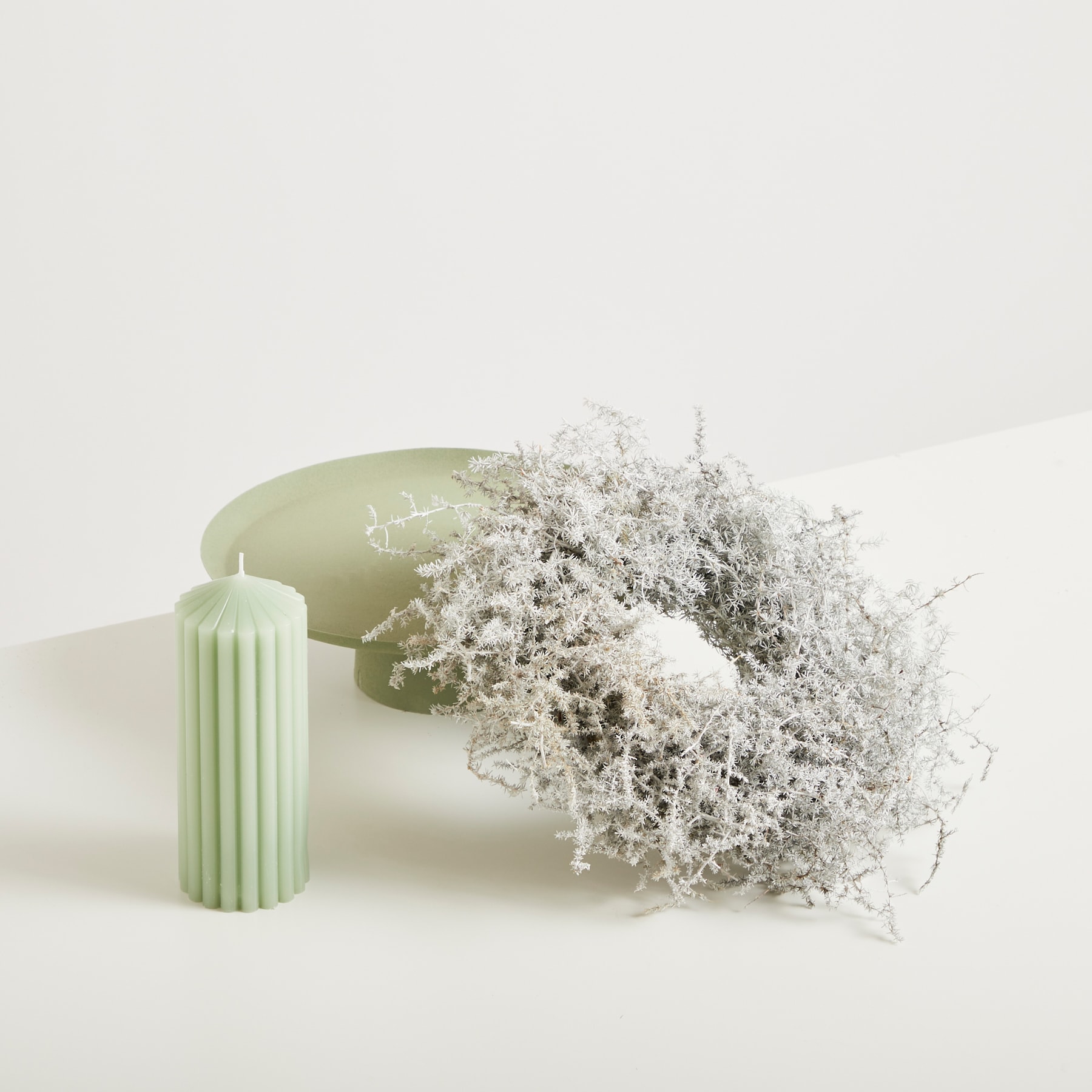 Auf einer schlichten weißen Oberfläche steht eine hellgrüne Keramikvase oder ein Behälter, aus dem ein zartes, hauchzartes Blumenarrangement hervorquillt.