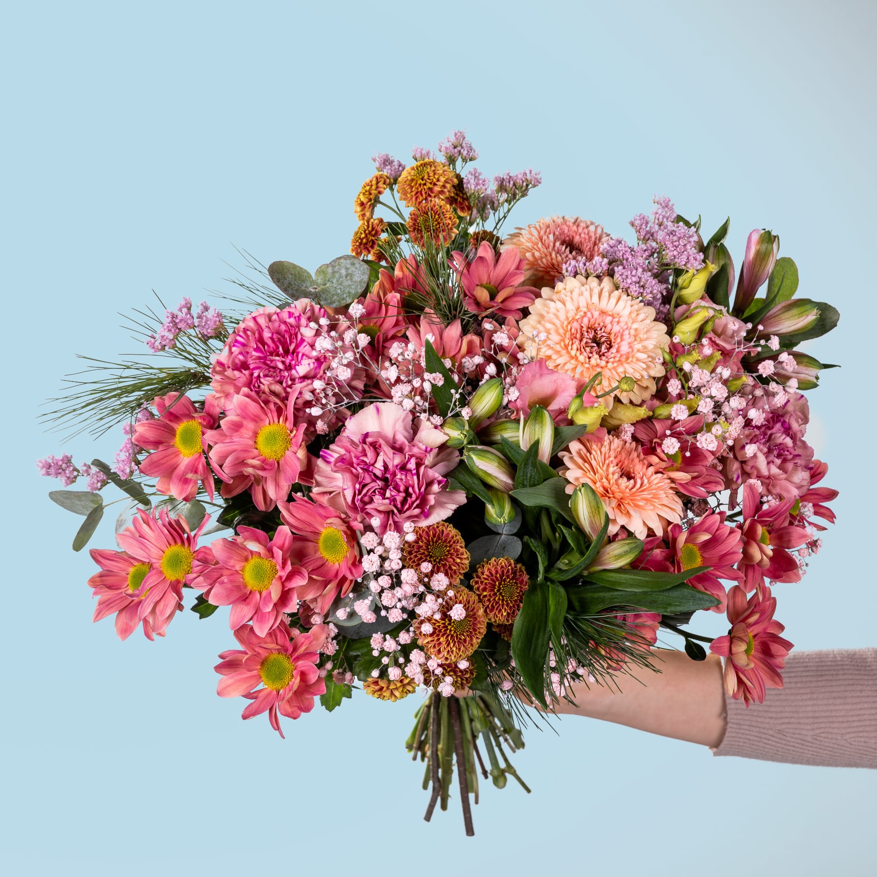 Ein farbenfroher Strauß verschiedener rosa, orangefarbener und gelber Blumen, darunter Dahlien, Chrysanthemen und andere Blüten, die eine Person vor einem schlichten blauen Hintergrund in den Händen hält.