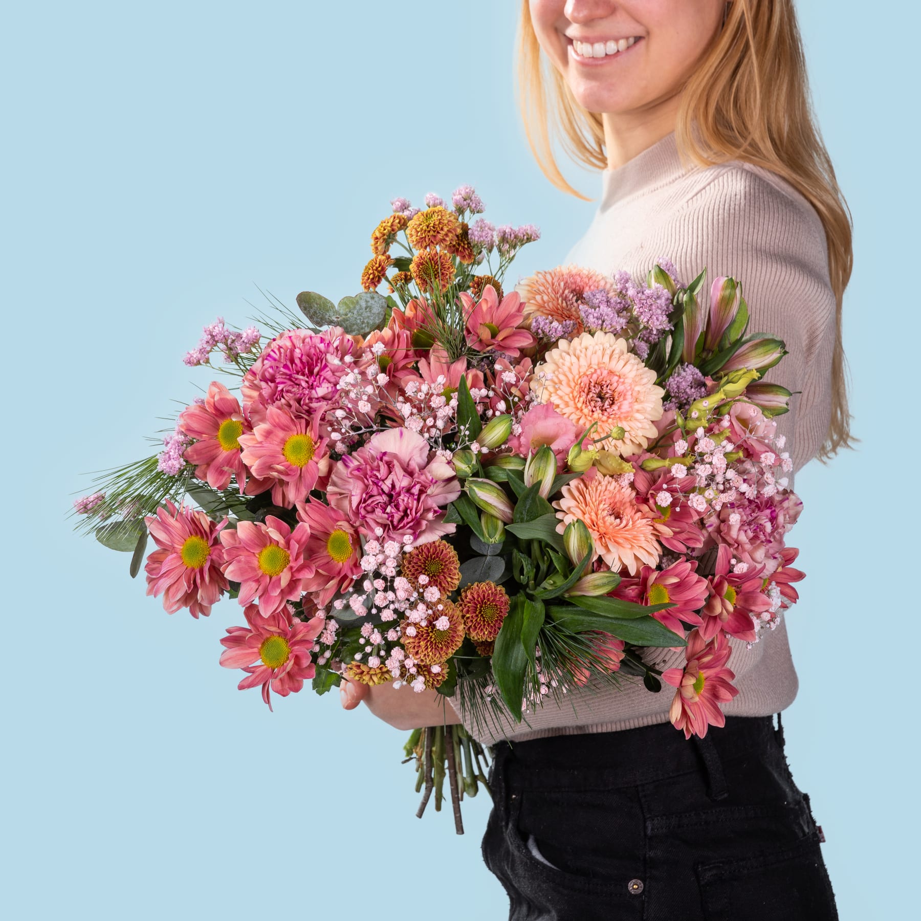 Eine lächelnde Frau hält vor einem schlichten blauen Hintergrund einen leuchtend bunten Strauß Blumen, darunter rosa und orangefarbene Dahlien.