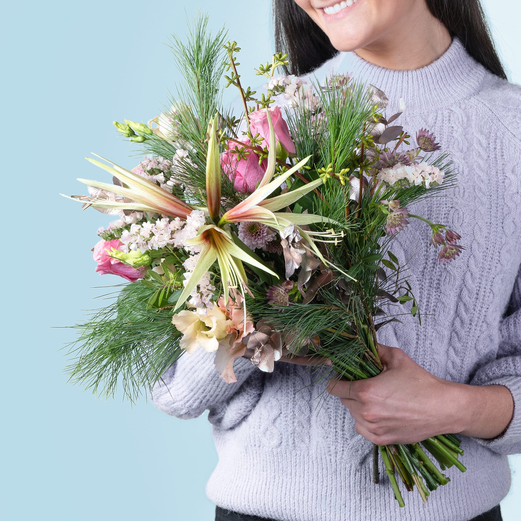 Eine Person in einem weißen Pullover hält einen großen, farbenfrohen Strauß verschiedener Blumen und Grünpflanzen vor einem hellblauen Hintergrund.