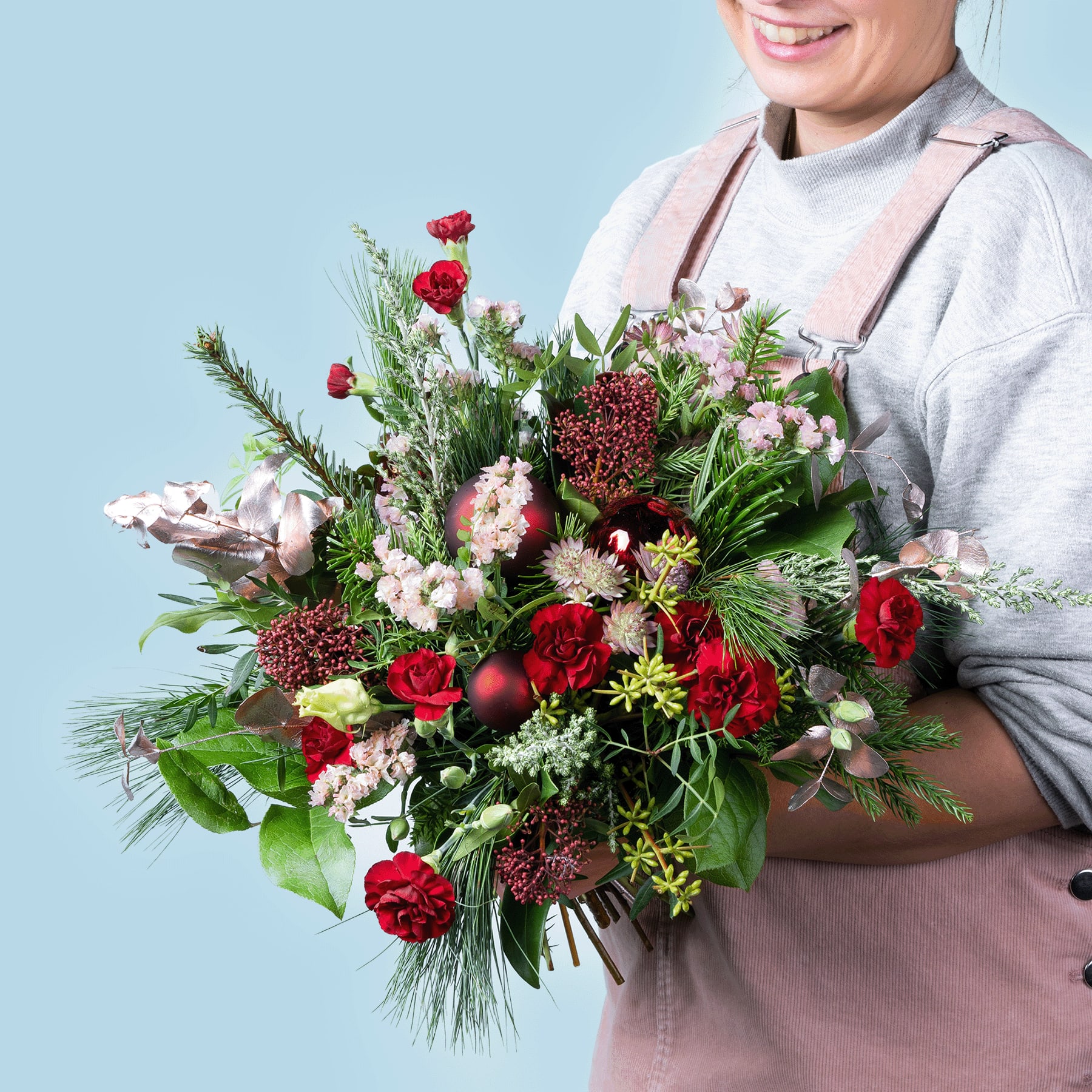Eine Person in einem rosa Pullover hält einen farbenfrohen Strauß aus roten Rosen, rosa Blumen und Grünpflanzen vor einem hellblauen Hintergrund.