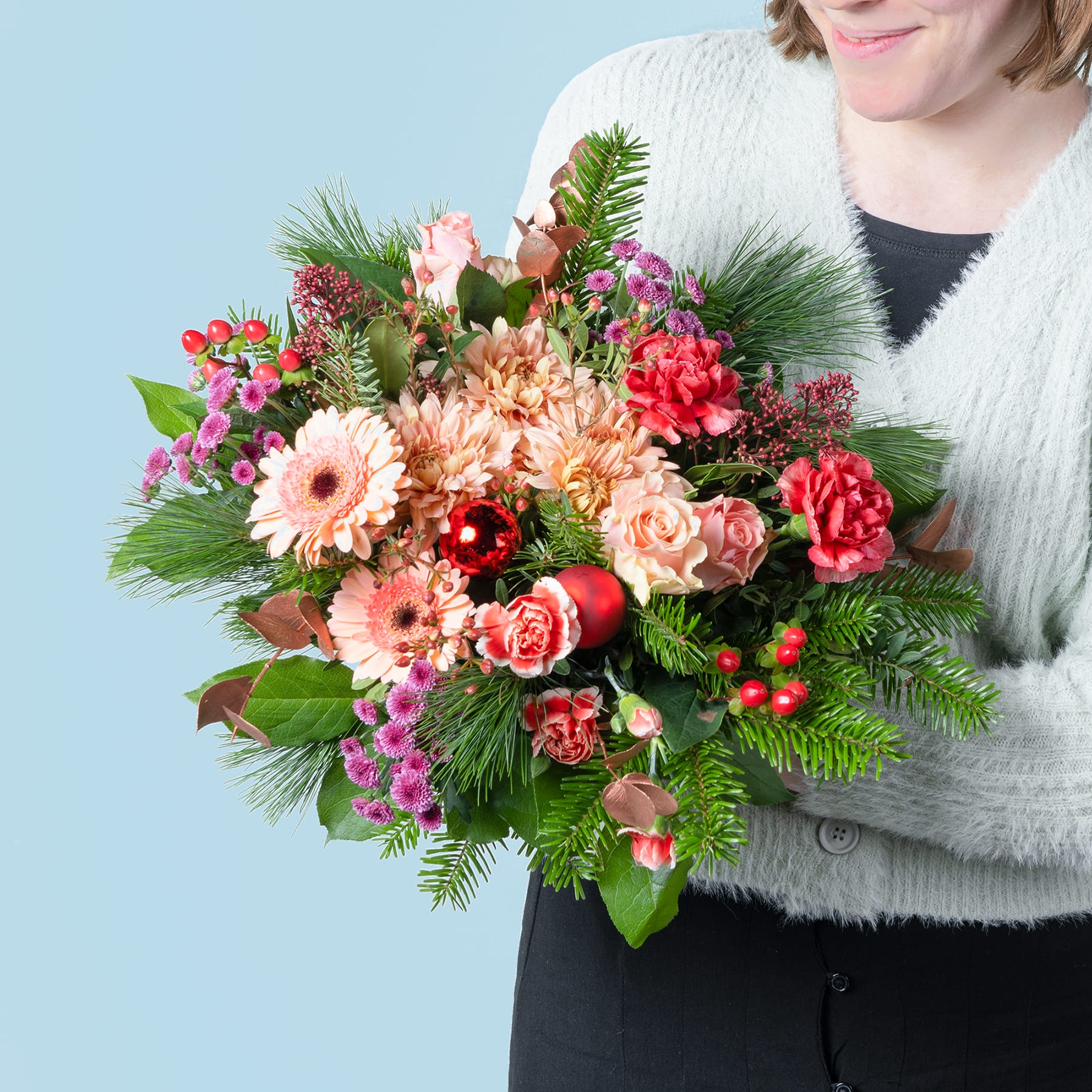 Ein leuchtender Strauß bunter Blumen, darunter rosa und orangefarbene Gerbera, rote Nelken und üppiges Grün, wird von einer Person gehalten, die einen weißen Pelzmantel vor einem hellblauen Hintergrund trägt.