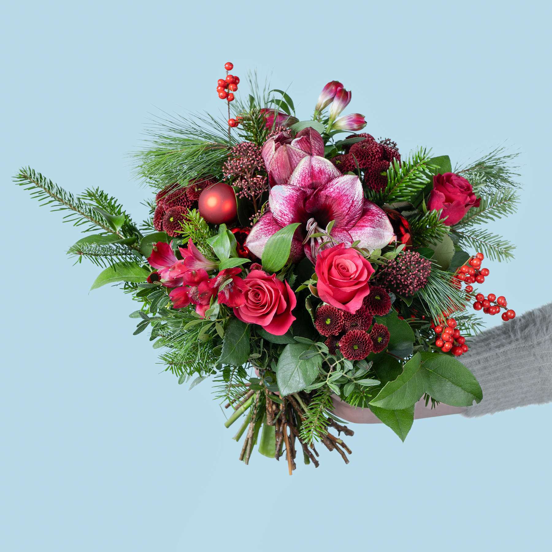 Ein leuchtender Strauß roter und rosa Blumen, darunter Rosen, Lilien und andere Grünpflanzen, vor einem hellblauen Hintergrund.