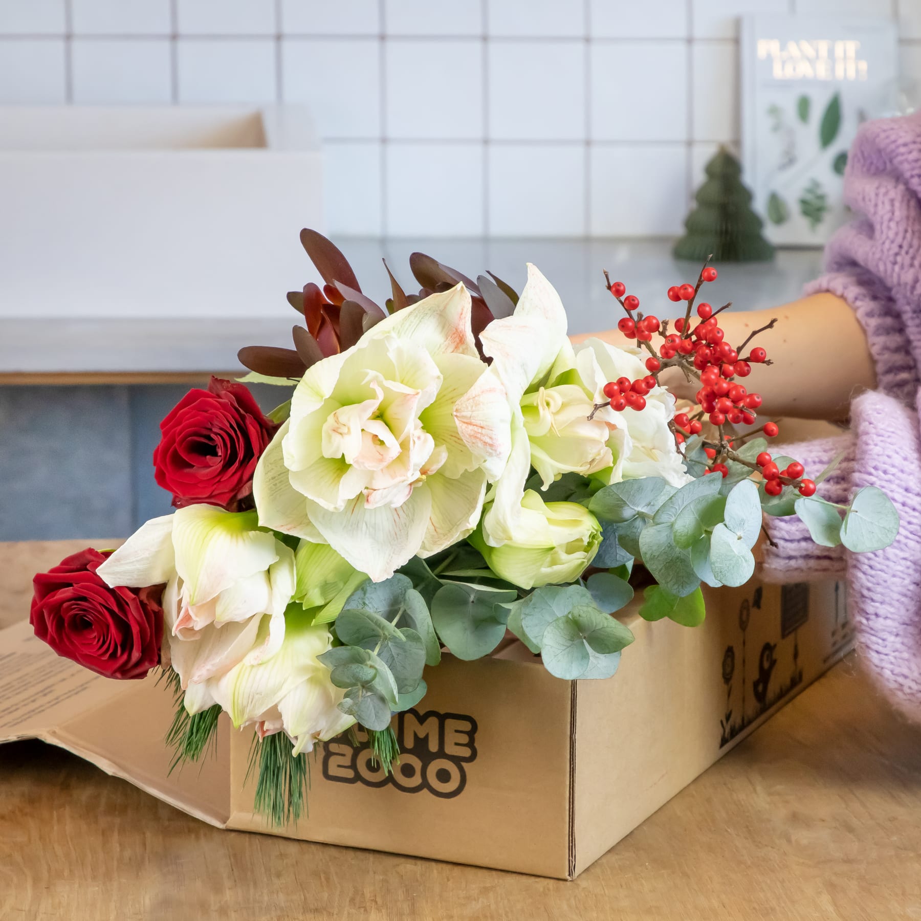 Ein bunter Blumenstrauß, darunter rote Rosen und weiße Hortensien, steht in einem Karton auf einer Holzoberfläche, im Hintergrund sind eine geflieste Wand und Dekorationsgegenstände zu sehen.