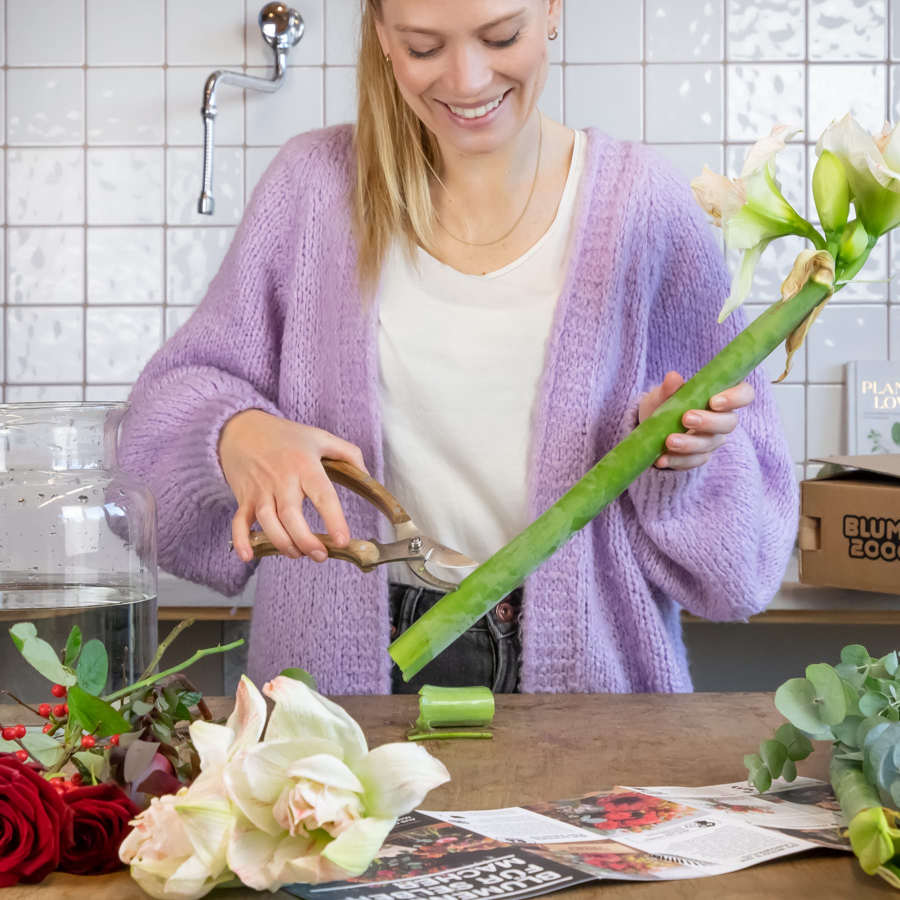 Eine lächelnde Frau in einer lila Strickjacke bereitet Blumen auf einer Küchentheke vor, umgeben von verschiedenen Pflanzen und einem Kochbuch.