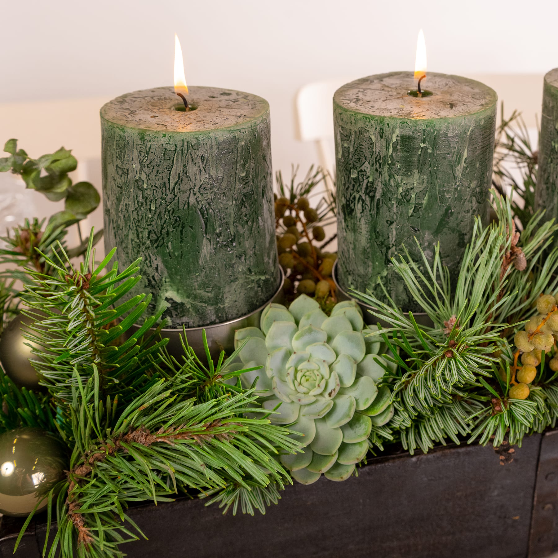 Das Bild zeigt ein festliches Weihnachtsarrangement mit zwei grünen Kerzen auf Holzscheiten, umgeben von verschiedenen Grünpflanzen wie Tannenzweigen, Sukkulenten und anderen dekorativen Elementen.
