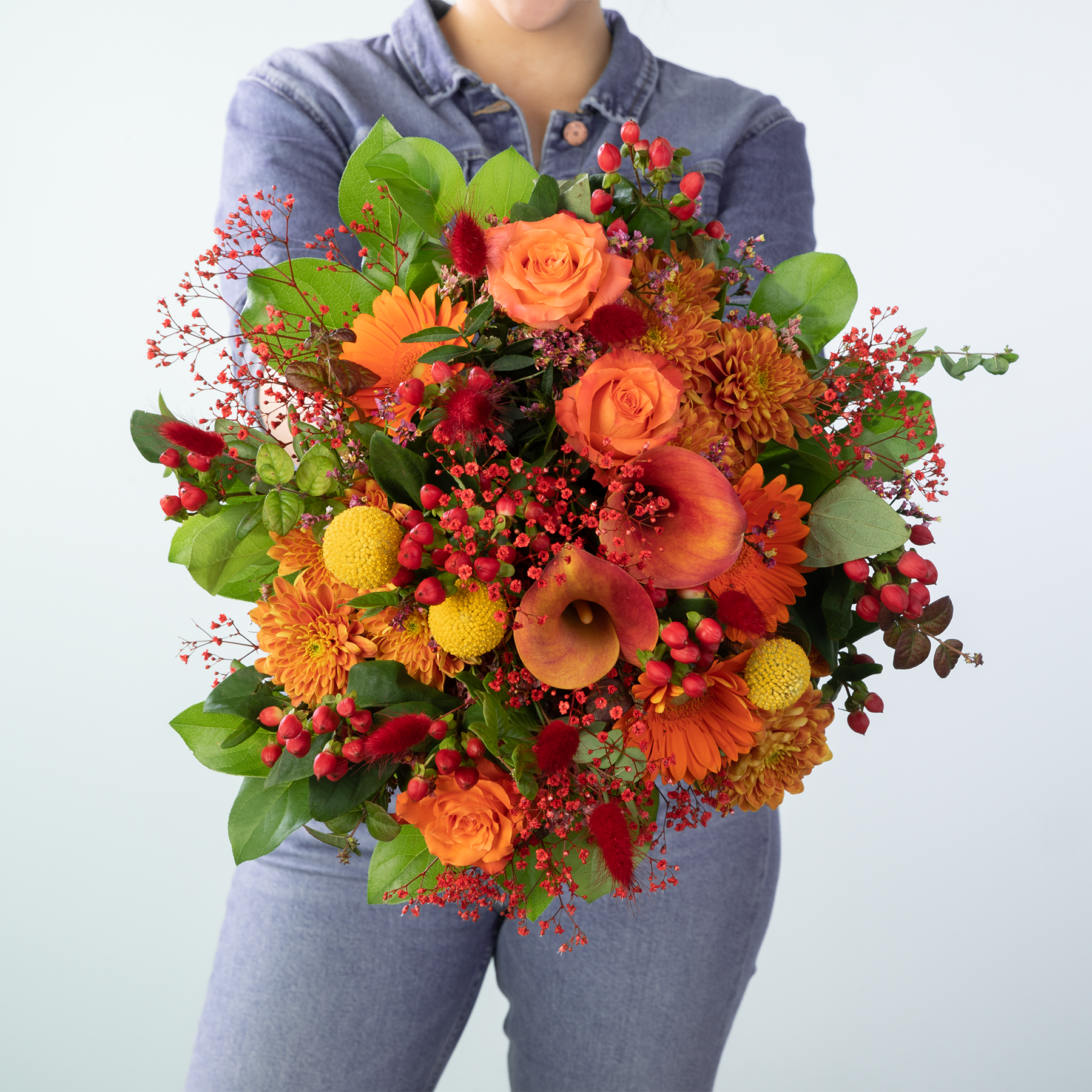 Ein farbenfroher Strauß herbstlicher Blumen, darunter orangefarbene Rosen, gelbe Chrysanthemen und rote Beeren, wird von einer Person gehalten, die ein graues Hemd vor einem schlichten weißen Hintergrund trägt.