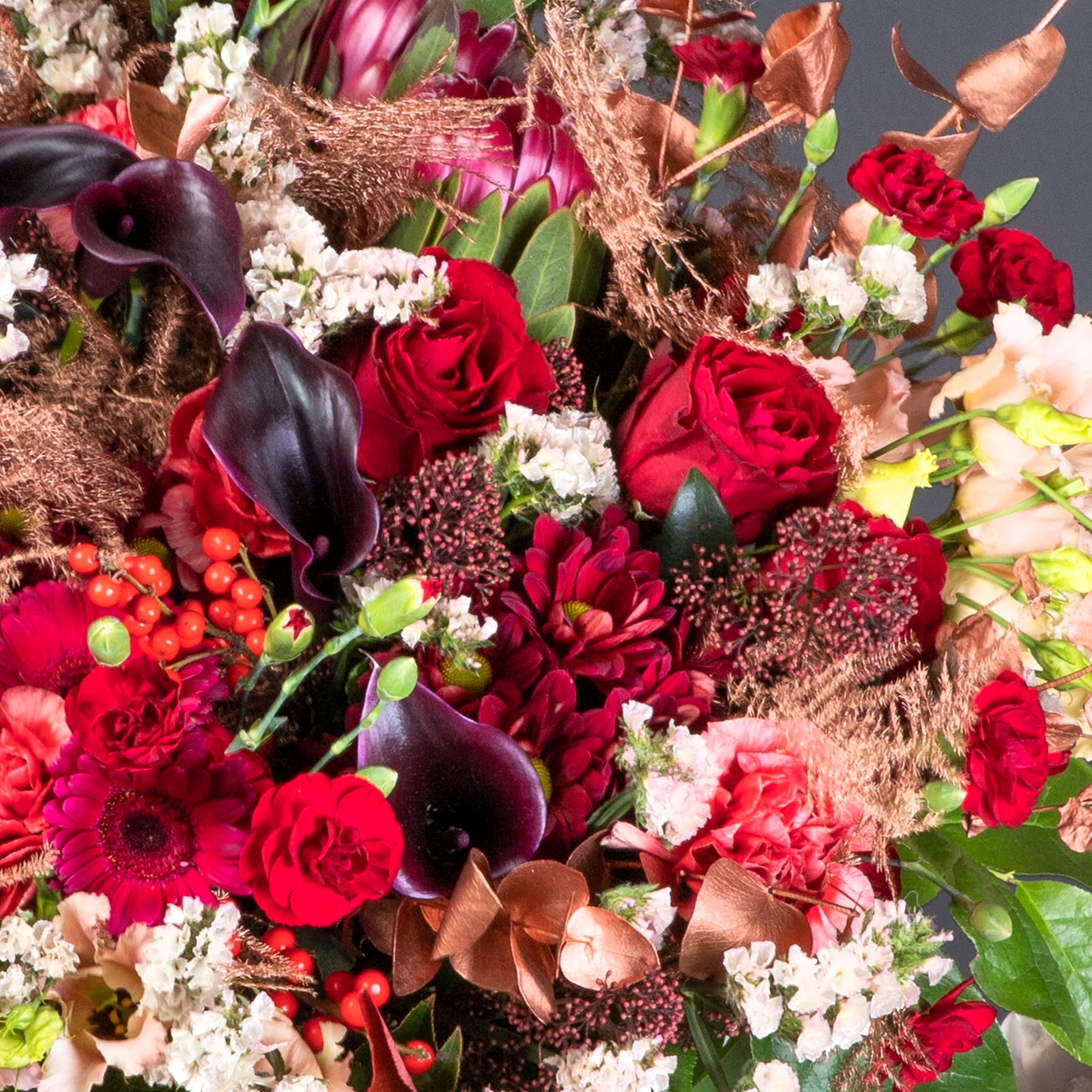 Ein lebendiger Strauß tiefroter Rosen, dunkelvioletter Callas und anderer üppiger, farbenfroher Blumen, arrangiert vor einem natürlichen, erdigen Hintergrund.