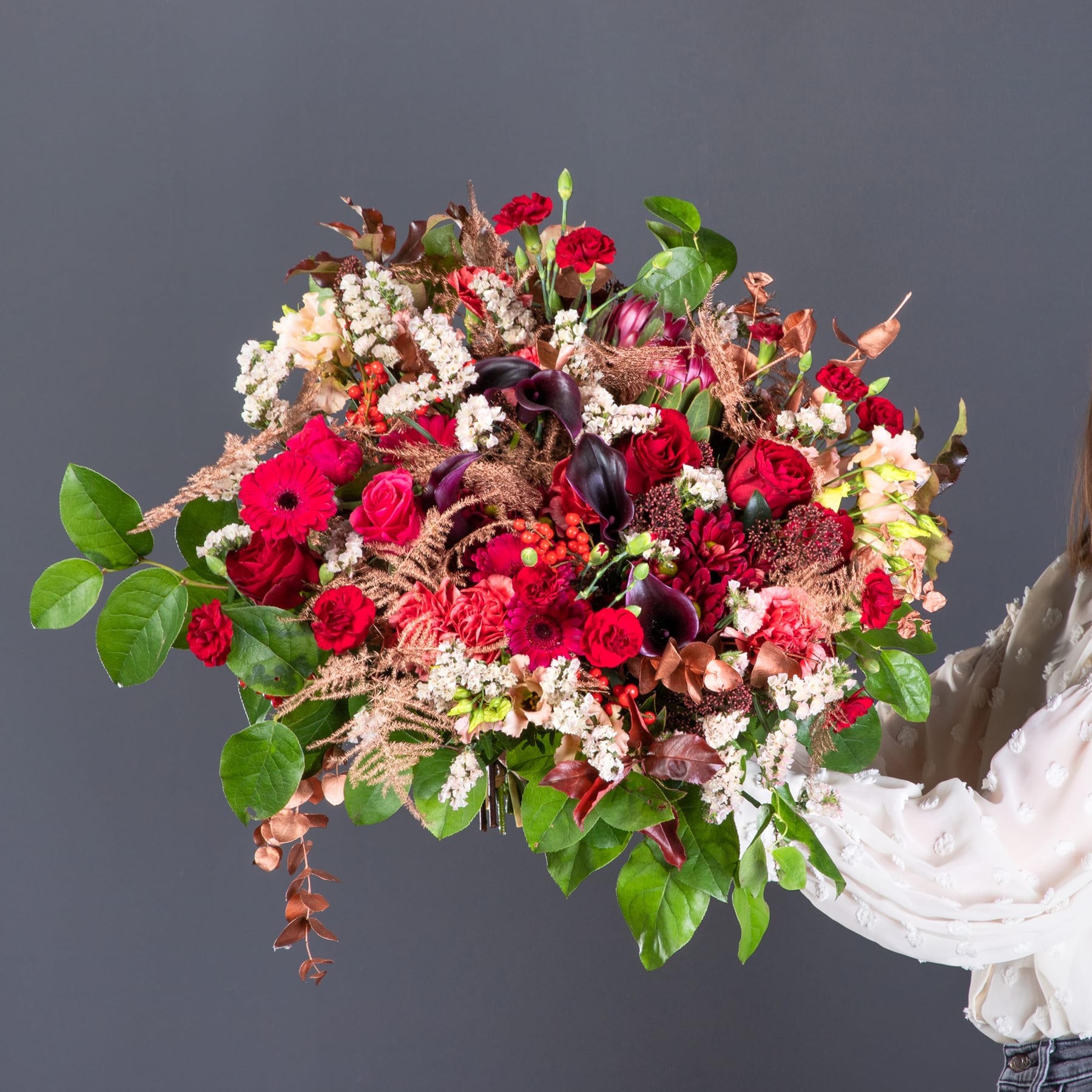 Ein lebendiger Strauß roter Rosen, Gerbera und anderer bunter Blumen, in rustikalem, natürlichem Stil vor einem schlichten grauen Hintergrund arrangiert.