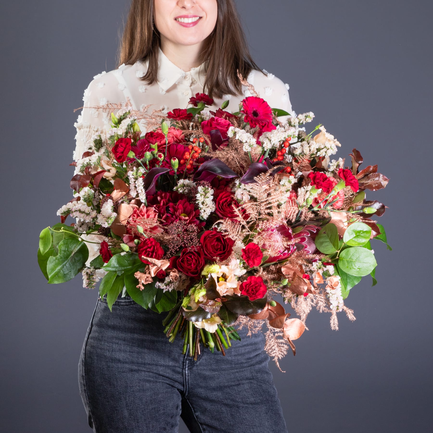 Eine junge Frau mit einem warmen Lächeln hält einen großen, leuchtenden Strauß aus roten Rosen, rosa Nelken und anderen bunten Blumen vor einem schlichten grauen Hintergrund.