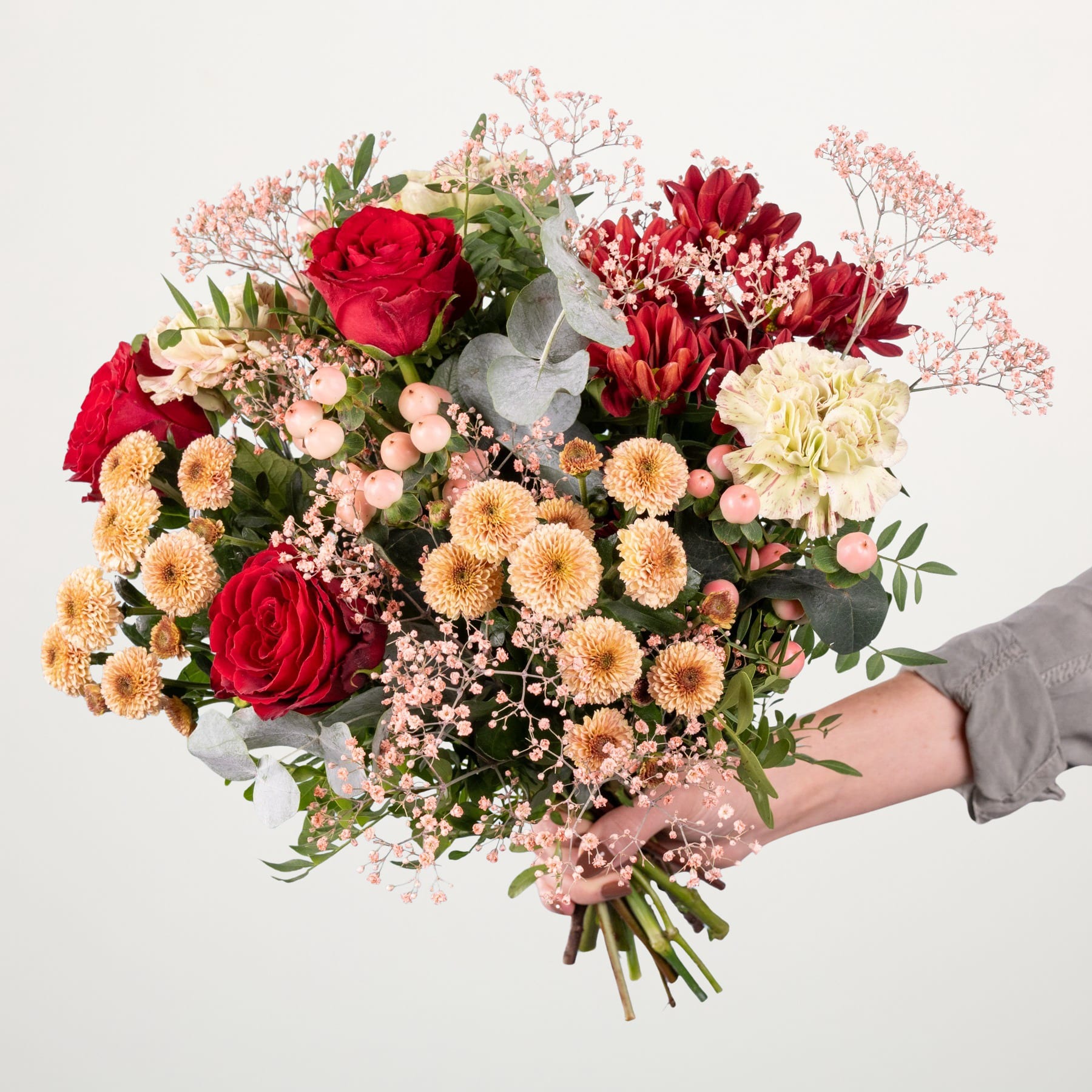 Ein leuchtender Strauß roter Rosen, rosa Chrysanthemen und anderer bunter Blumen, den eine Person vor einem schlichten weißen Hintergrund in den Händen hält.