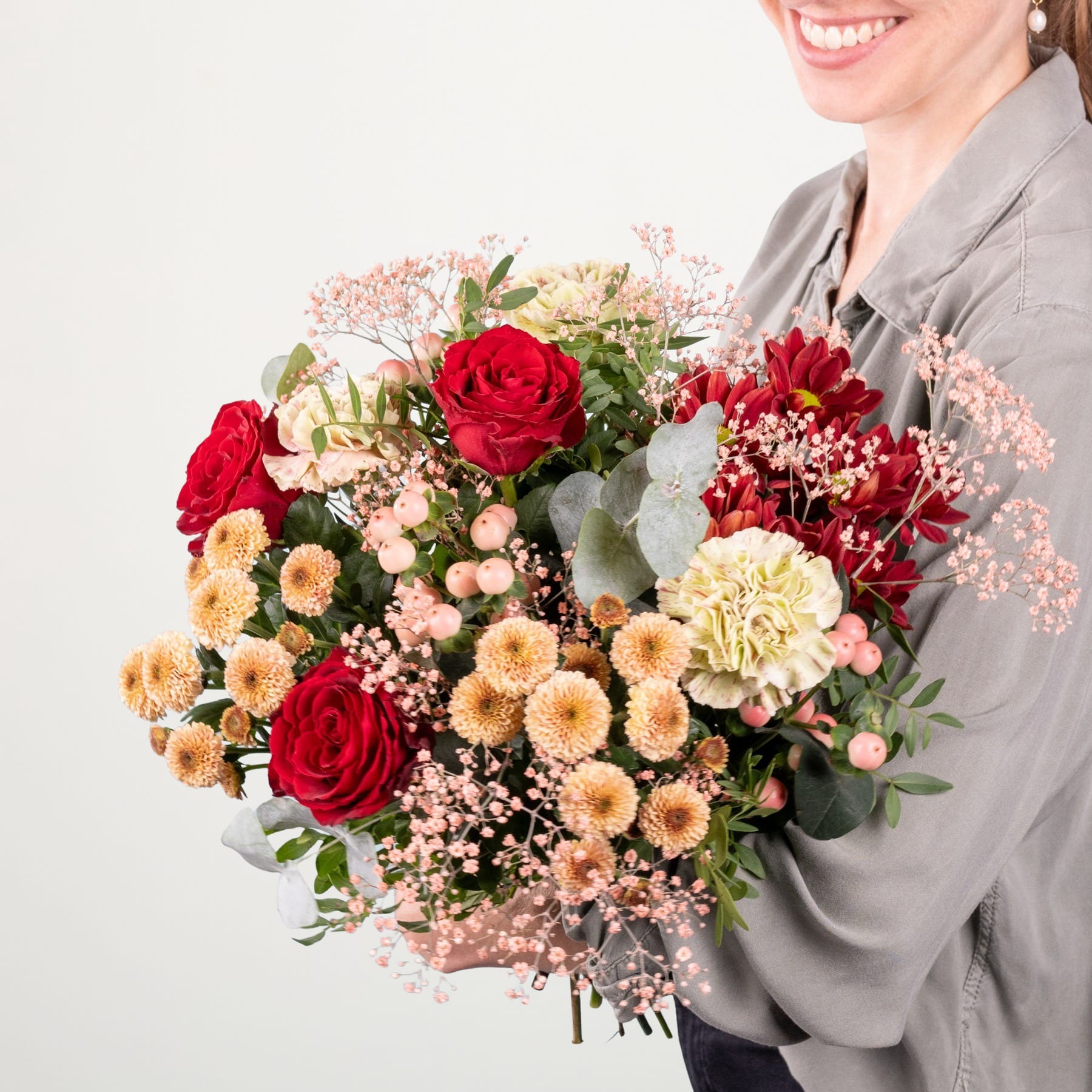 Ein leuchtender Strauß roter Rosen, gelber Chrysanthemen und anderer bunter Blumen wird von einer lächelnden Person in einem grauen Hemd vor einem schlichten Hintergrund gehalten.