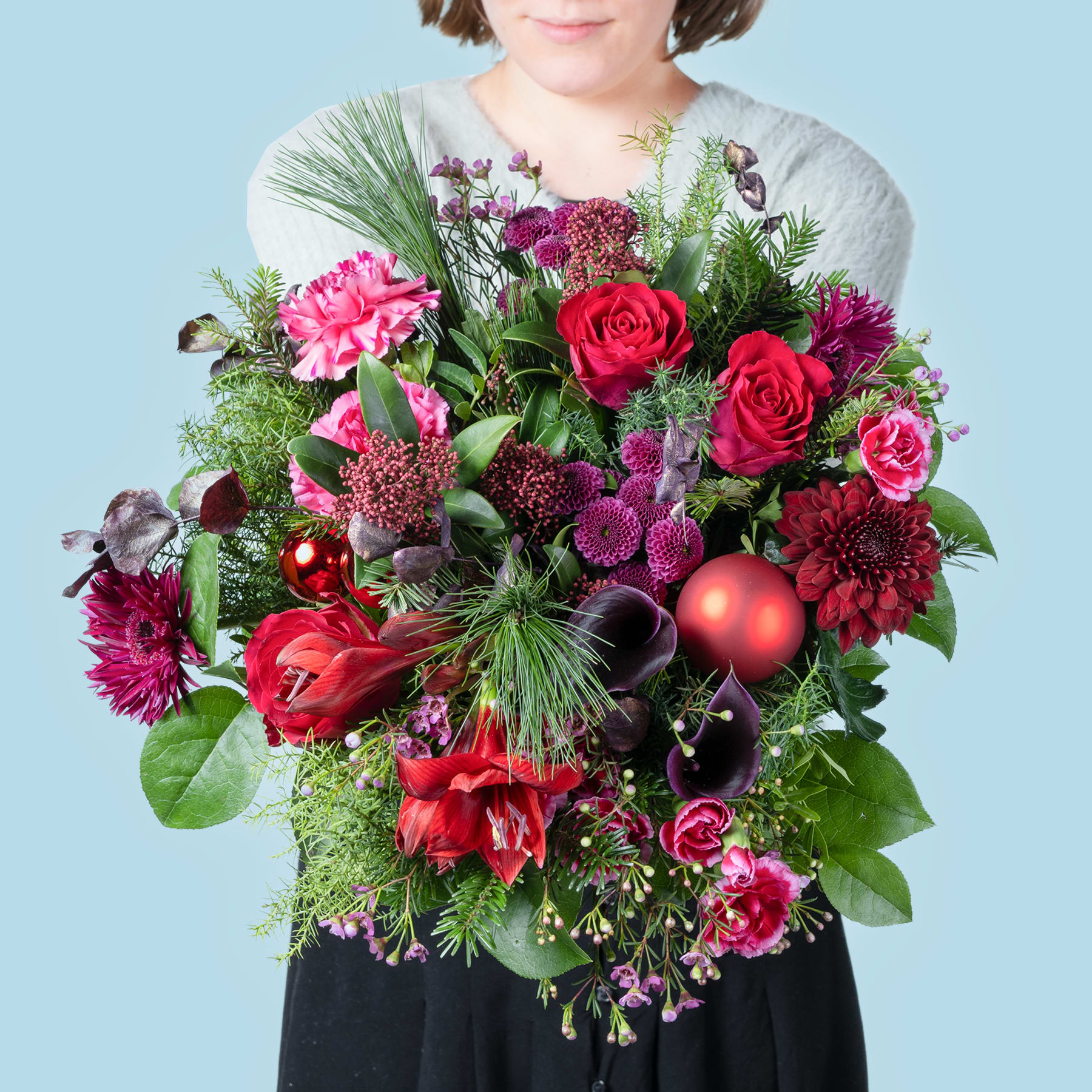 Ein leuchtender Strauß roter Rosen, rosa Pfingstrosen und verschiedener anderer bunter Blumen wird von einer Person gehalten, die einen grauen Pullover vor einem hellblauen Hintergrund trägt.