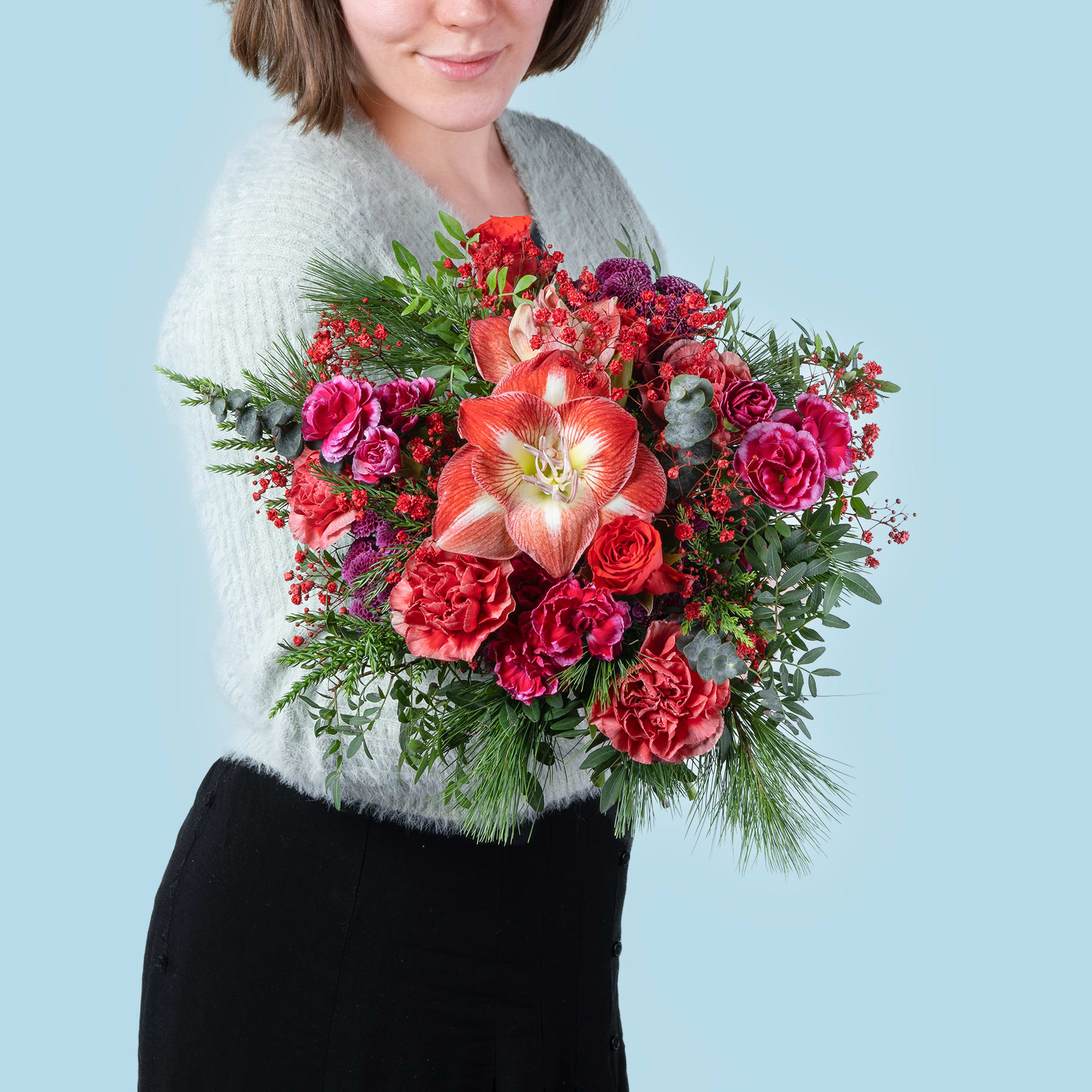 Eine Frau in einem grauen Pullover hält vor einem hellblauen Hintergrund einen farbenfrohen Strauß roter und rosa Blumen, darunter Rosen und Lilien.