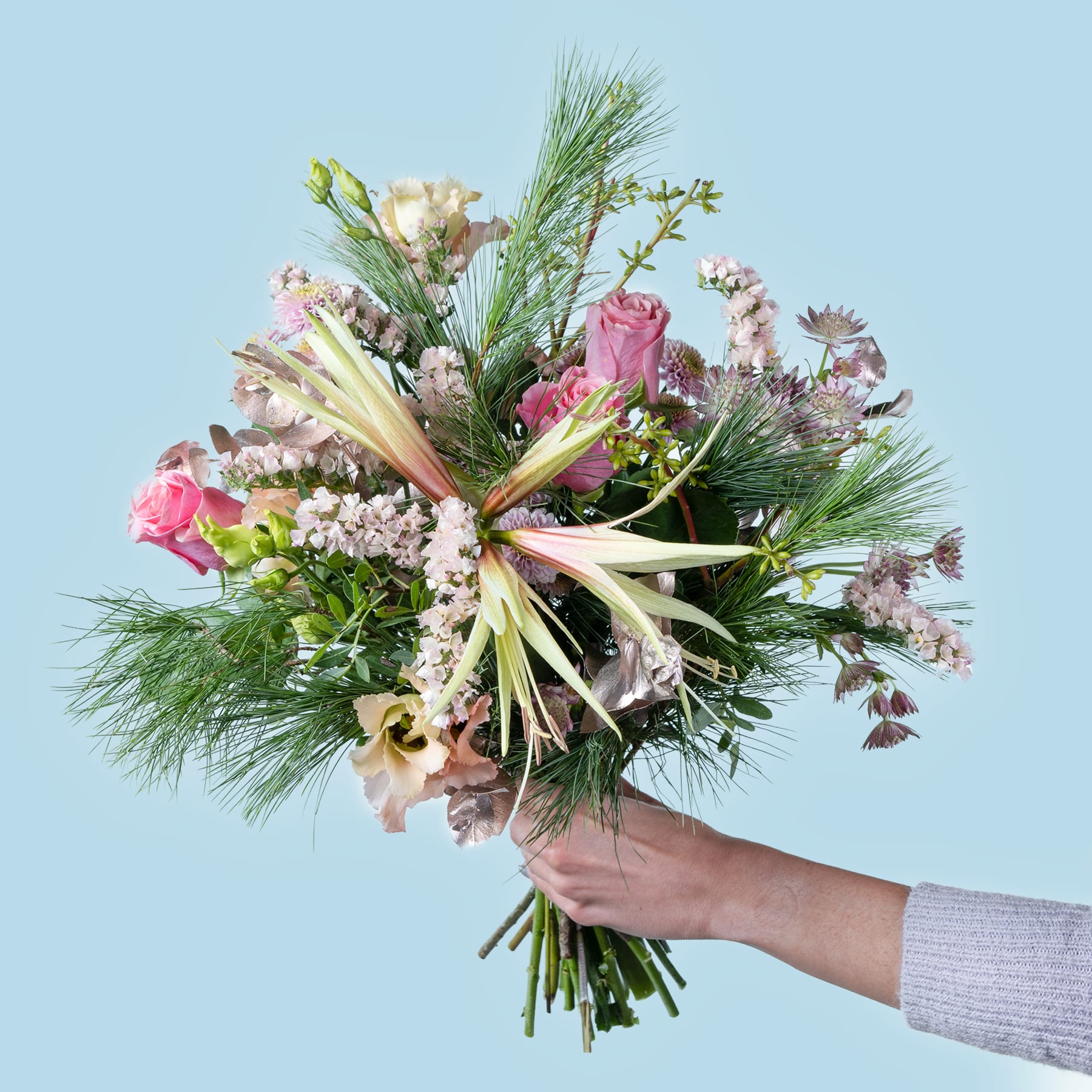 Ein farbenfroher Strauß verschiedener Blumen, darunter rosa Rosen, Tannenzweige und anderes Grün, wird vor einem hellblauen Hintergrund in der Hand einer Person gehalten.