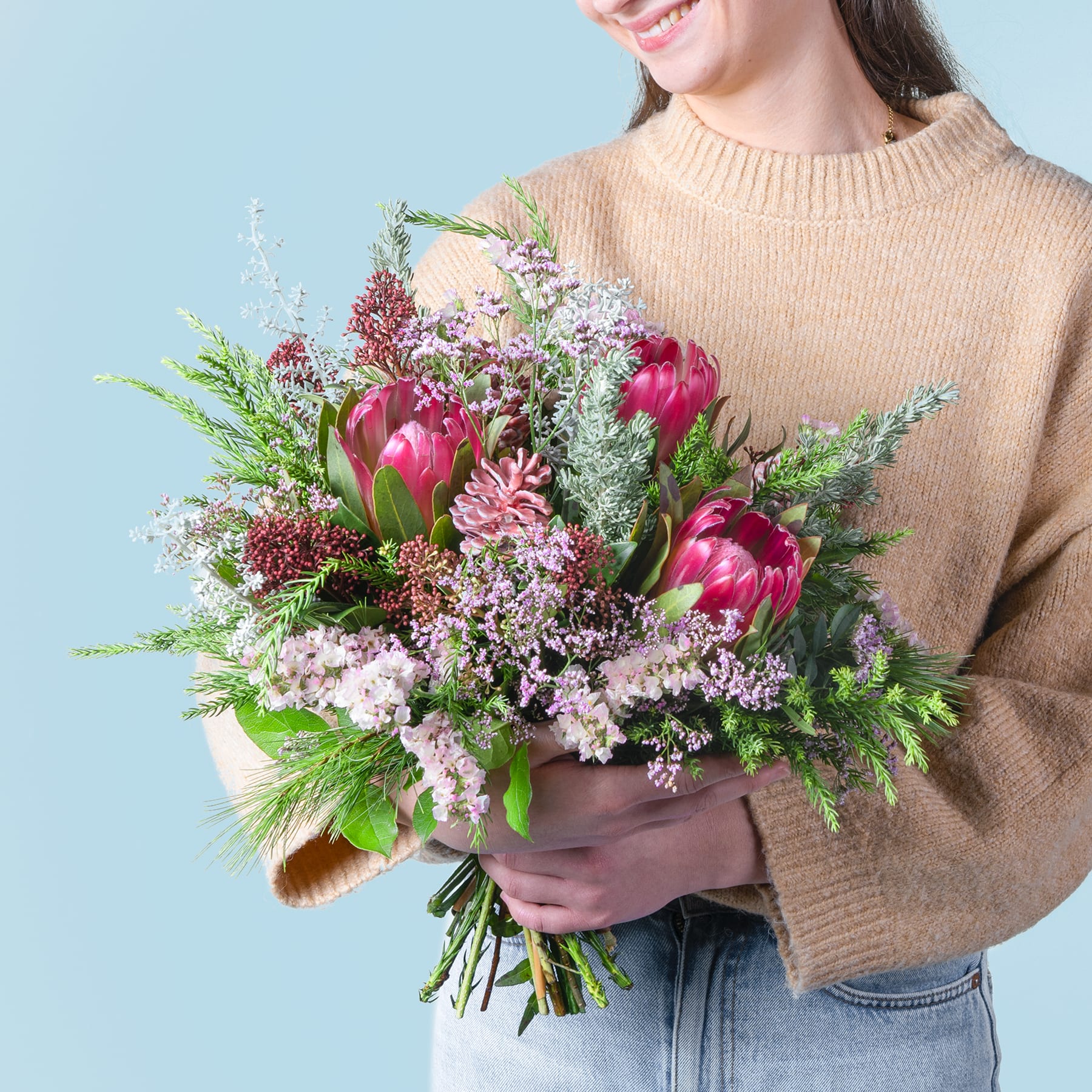 Eine Frau in einem beigen Pullover hält einen farbenfrohen Strauß aus rosa Protea-Blüten, violetten und weißen Wildblumen und üppigem Grün vor einem schlichten blauen Hintergrund.