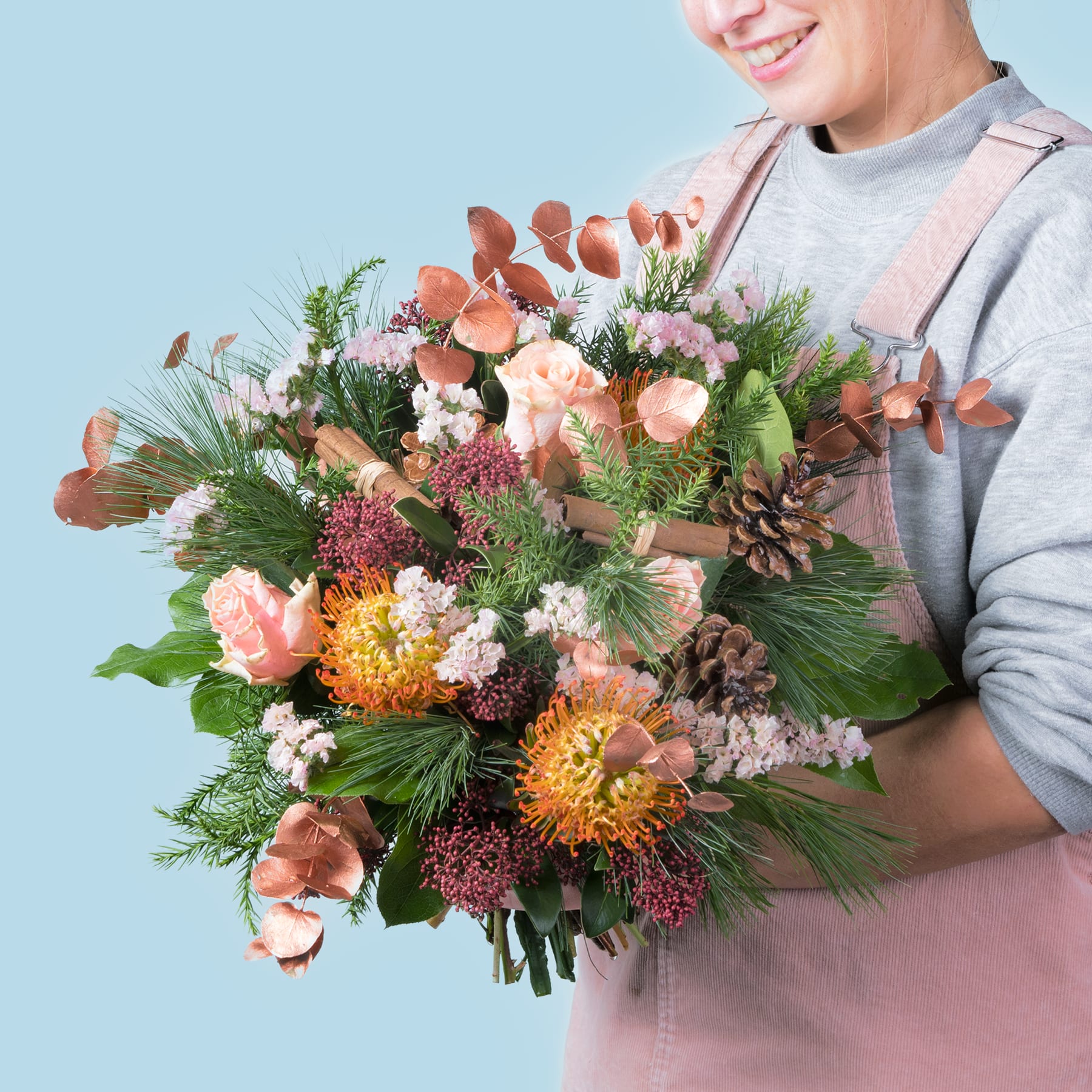 Ein farbenfroher Strauß verschiedener Blumen, darunter Rosen, Chrysanthemen und Tannenzweige, wird von einer Person gehalten, die einen hellen Pullover vor einem schlichten blauen Hintergrund trägt.