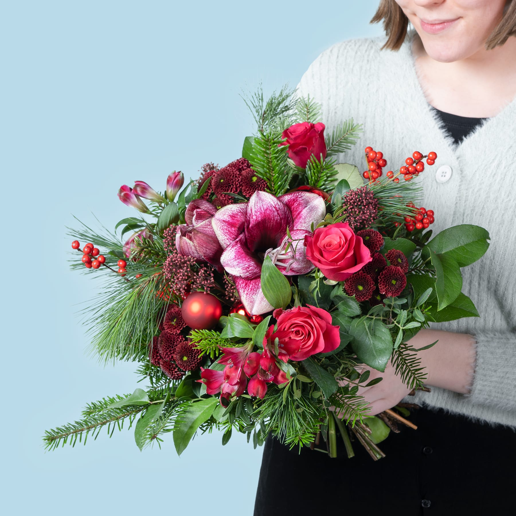 Ein leuchtender Strauß roter und rosa Blumen, darunter Rosen, Orchideen und Beeren, gehalten von einer Person in einem weißen Pullover vor einem schlichten blauen Hintergrund.
