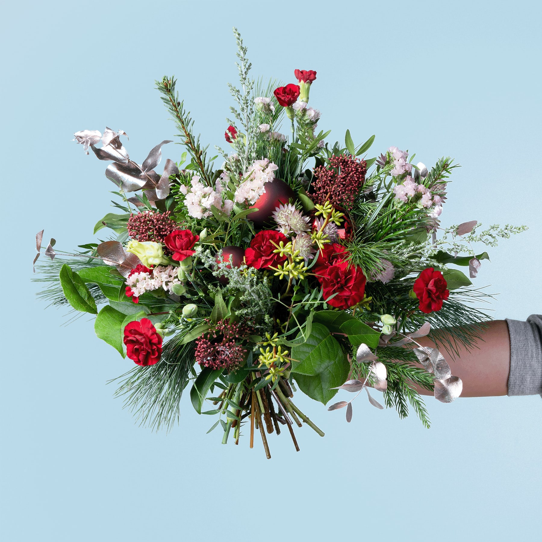 Ein farbenfroher Strauß verschiedener Blumen, darunter rote Rosen, weiße Gänseblümchen und Grünpflanzen, wird vor einem schlichten blauen Hintergrund in der Hand einer Person gehalten.