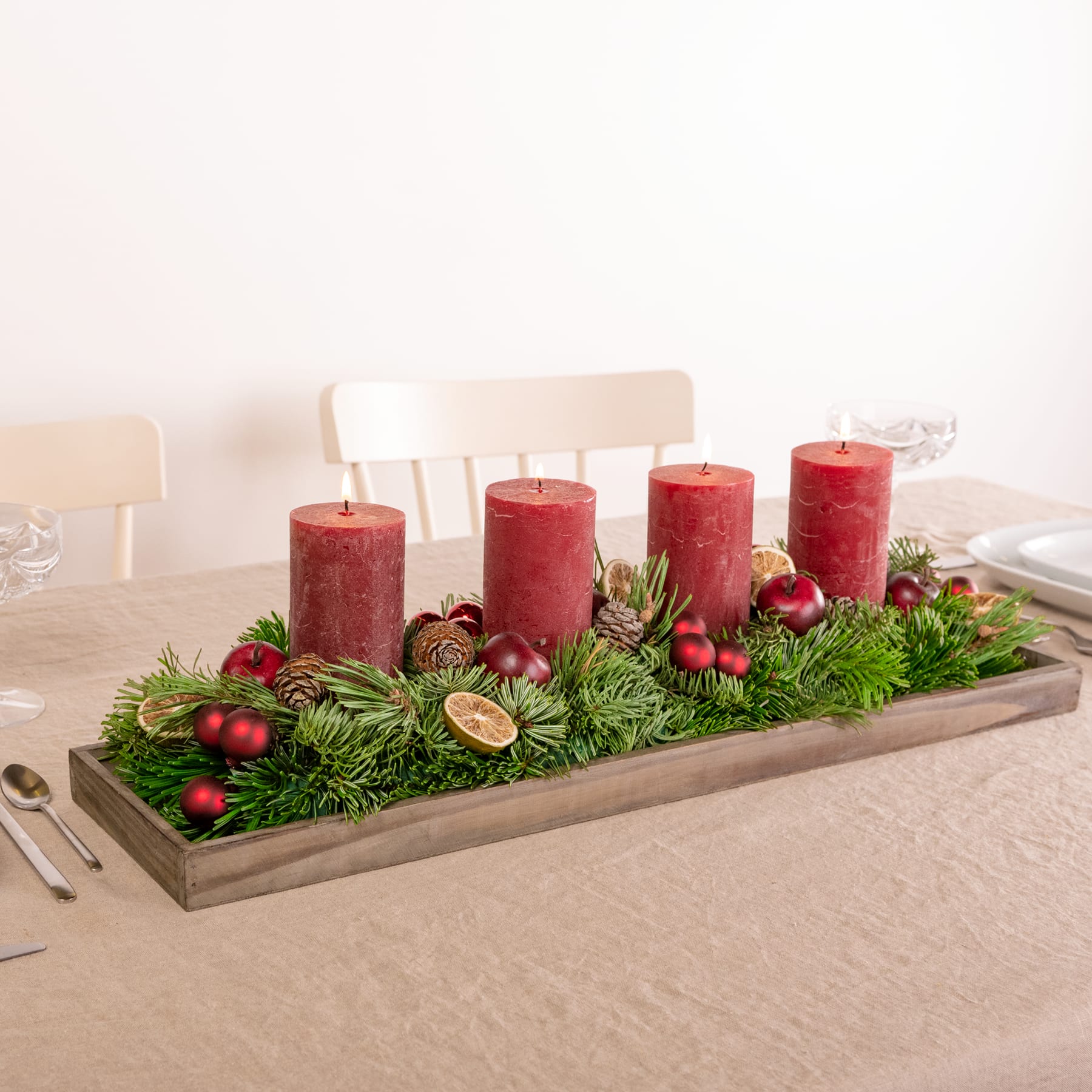 Ein Holztablett mit vier roten Kerzen, umgeben von einem festlichen Arrangement aus Tannenzweigen, Beeren und anderen Naturelementen, auf einem hellen Tisch vor einem schlichten Hintergrund.