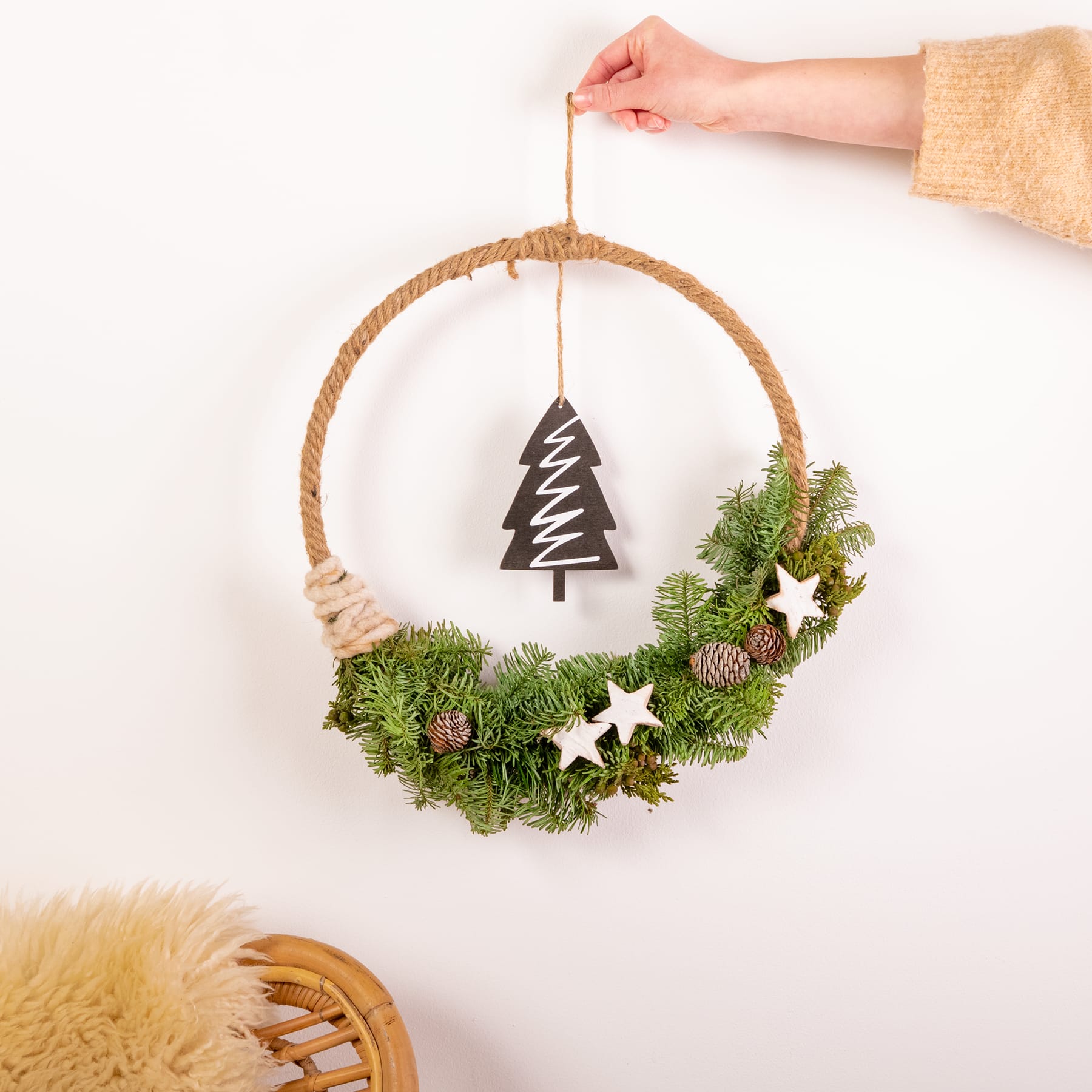 Eine Hand hängt einen dekorativen Weihnachtskranz aus immergrünen Zweigen, Sternen und einem hölzernen Baumschmuck an eine weiße Wand.