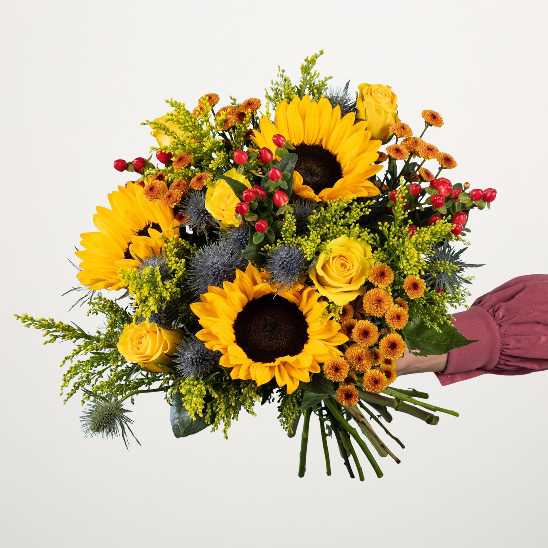 Ein farbenfroher Strauß aus Sonnenblumen, Rosen und anderen herbstlichen Blumen, der vor einem schlichten weißen Hintergrund in den Händen einer Person gehalten wird.
