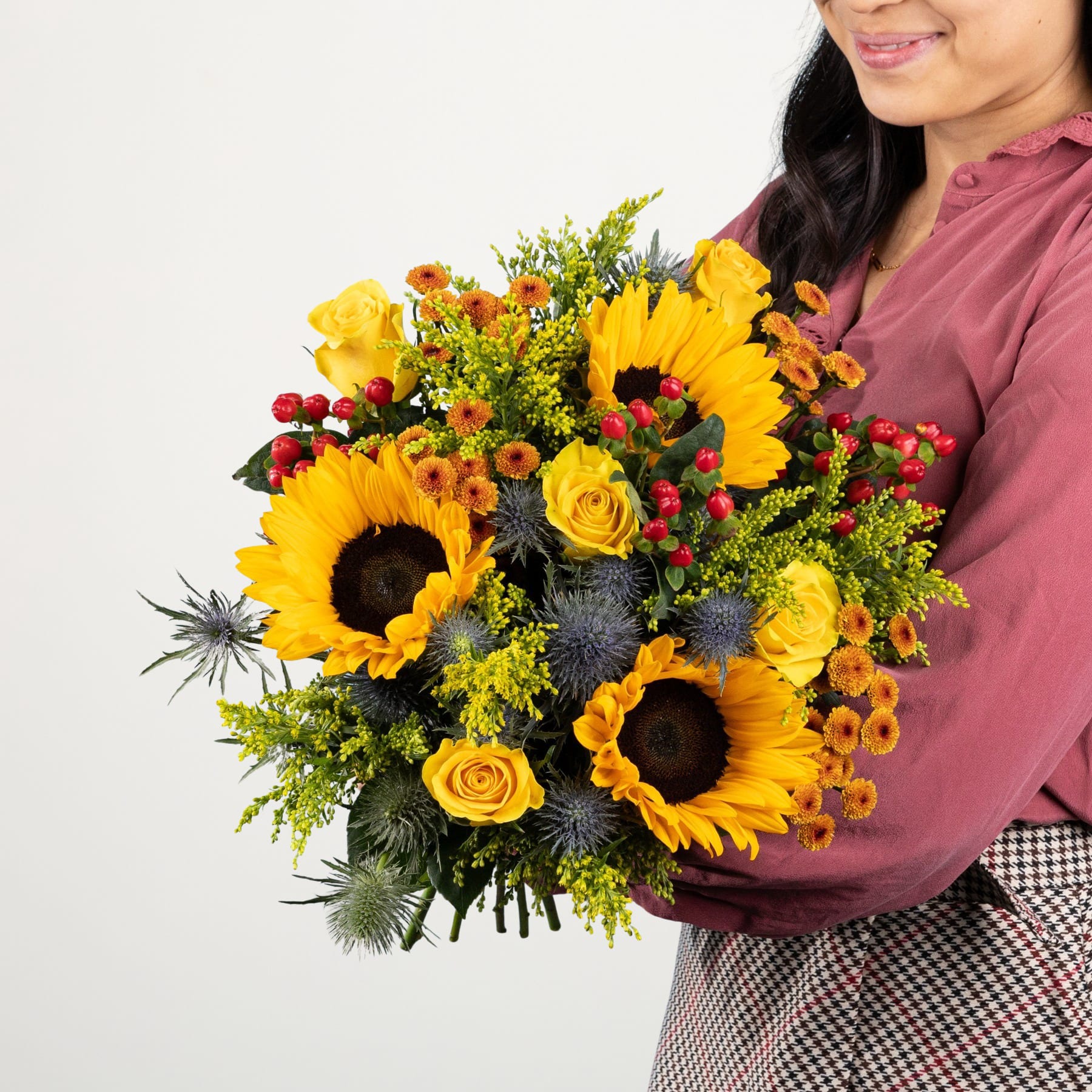Ein farbenfroher Strauß Sonnenblumen, Rosen und anderer herbstlicher Blumen wird von einer lächelnden Frau in einem rosa Hemd und einem karierten Rock vor einem schlichten weißen Hintergrund gehalten.