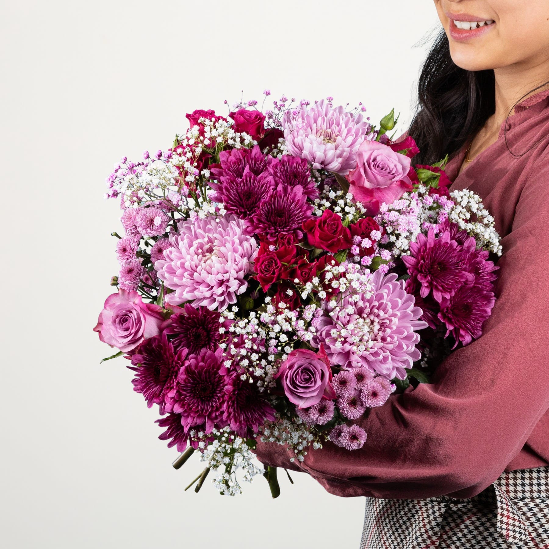 Ein farbenfroher Strauß rosa und violetter Blumen, darunter Rosen, Chrysanthemen und Schleierkraut, wird von einer Person gehalten, die einen burgunderfarbenen Pullover vor einem schlichten Hintergrund trägt.