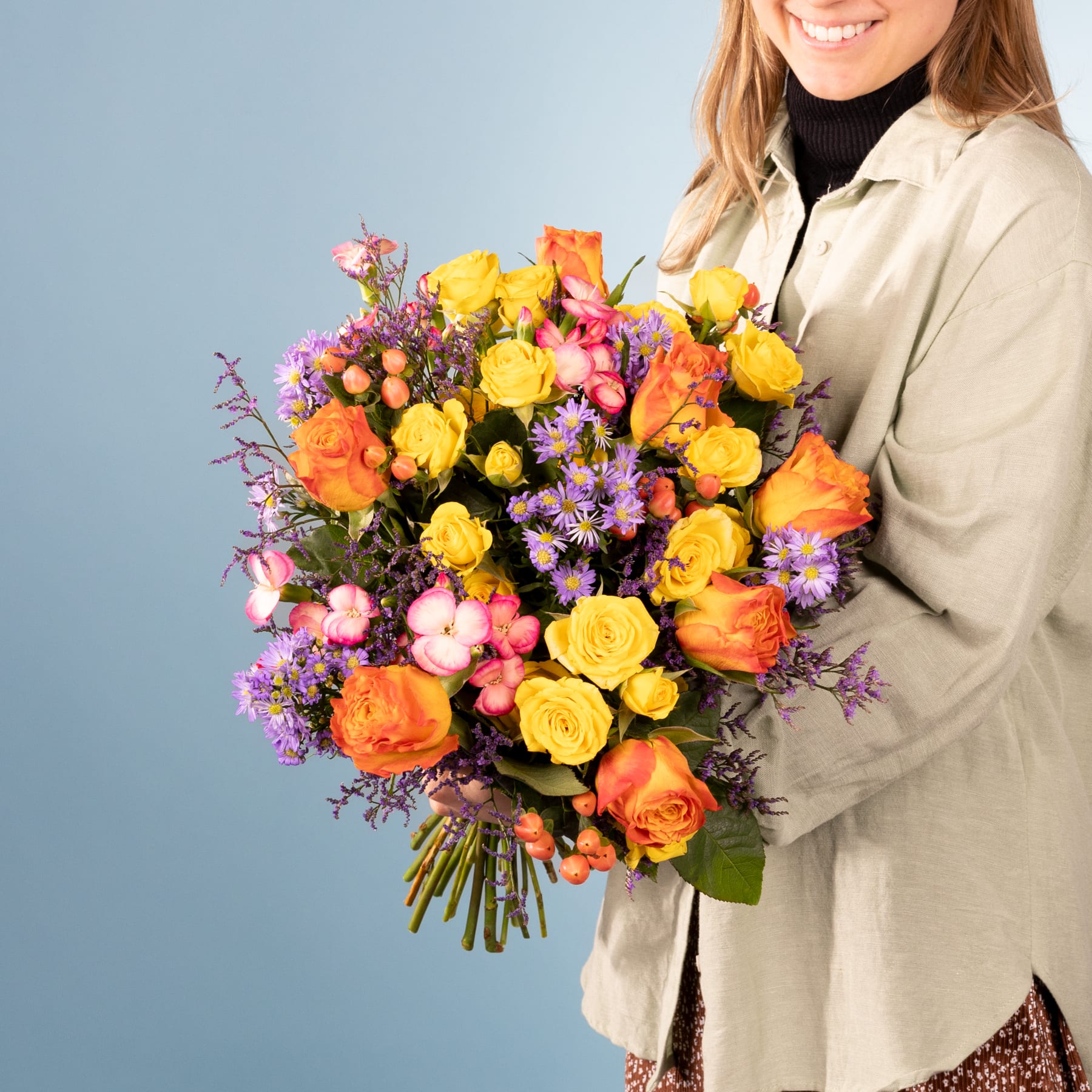 Eine lächelnde Frau in einem beigen Mantel hält einen farbenfrohen Strauß aus gelben und orangefarbenen Rosen, lila Gänseblümchen und anderen bunten Blumen vor einem schlichten grauen Hintergrund.