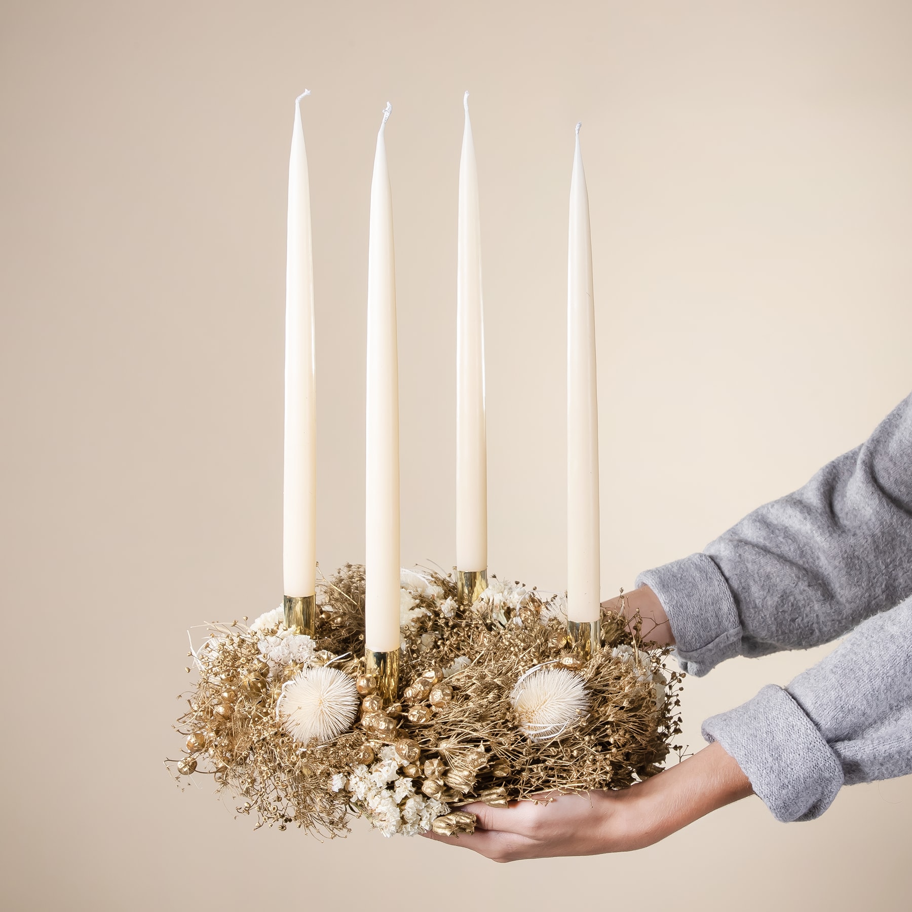 Ein kranzartiges Arrangement aus getrockneten Blumen und Blättern, geschmückt mit weißen Kerzen, wird vor einem neutralen Hintergrund in den Händen einer Person gehalten.