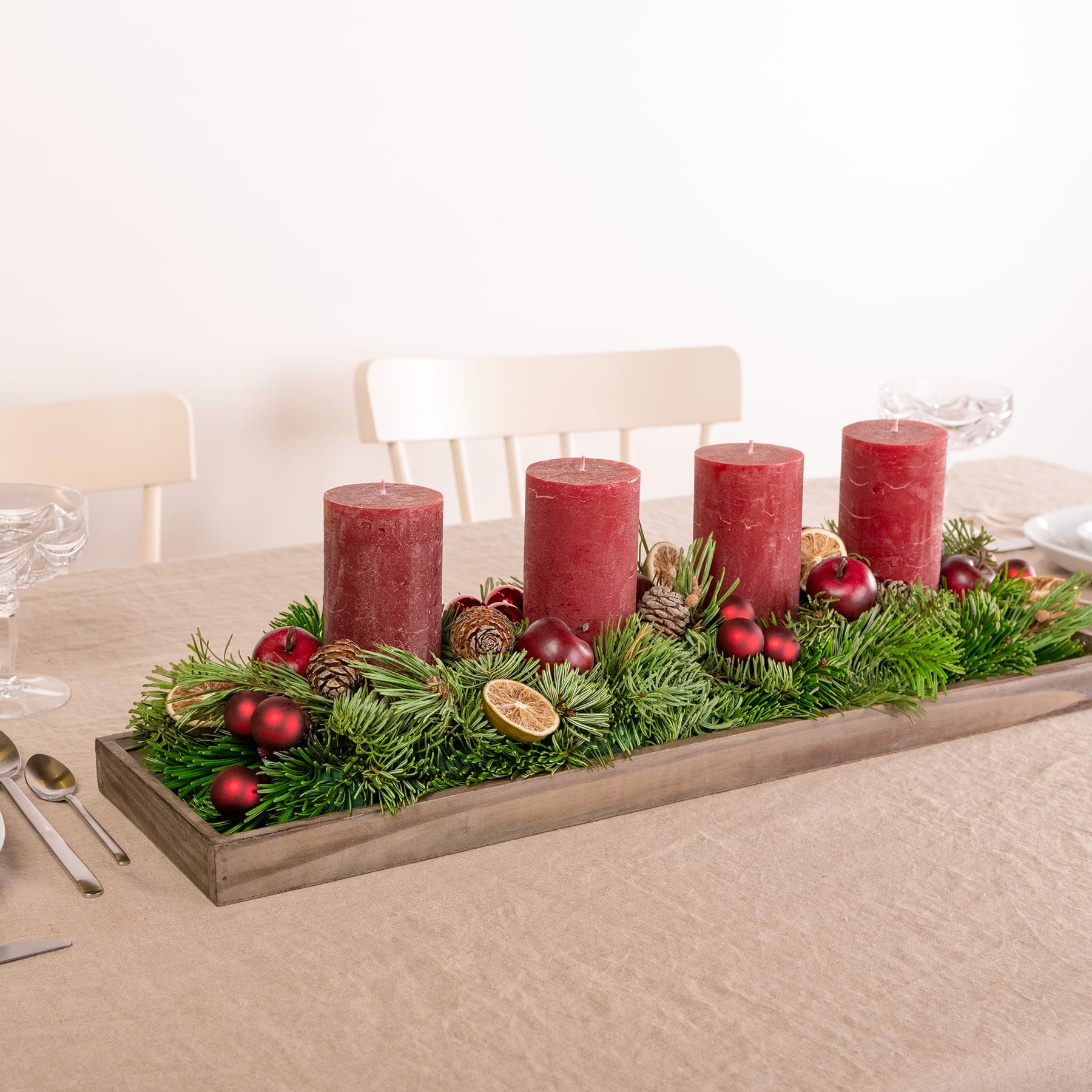 Ein Holztablett mit vier roten Kerzen, Tannenzweigen und verschiedenen Weihnachtsornamenten und -dekorationen vor einem schlichten Hintergrund.