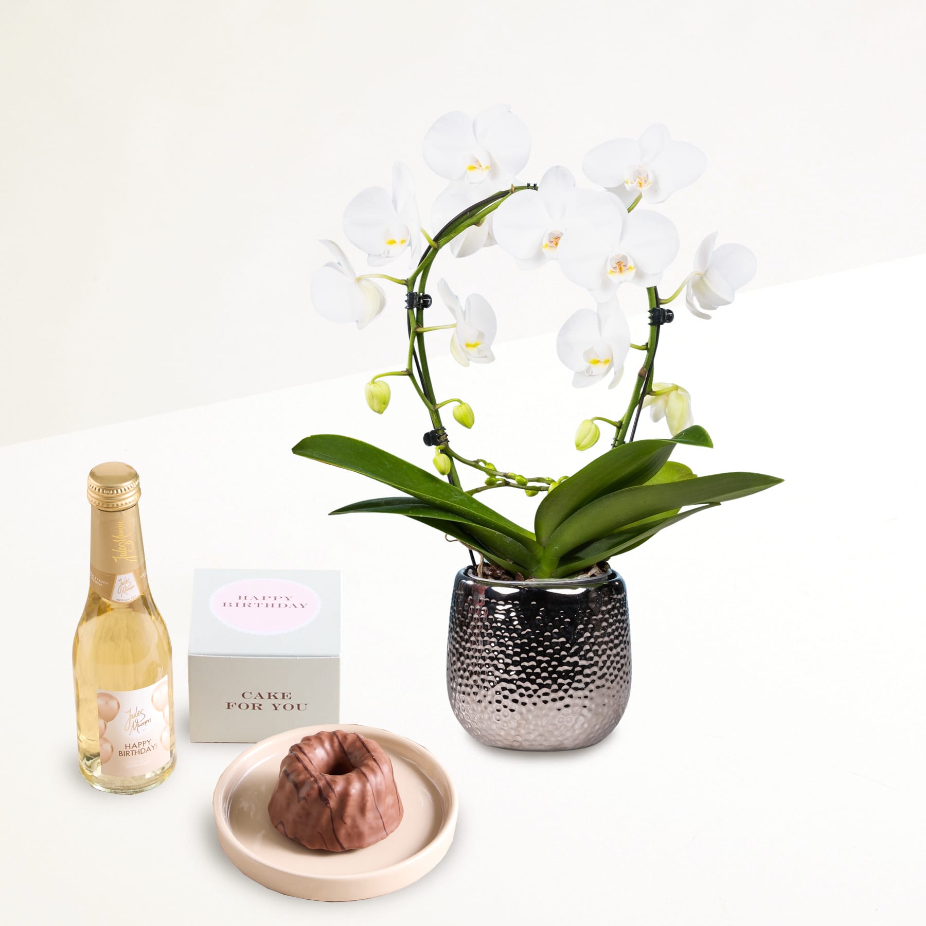 Eine weiße Orchidee im Topf, eine Flasche Wein und ein kleines Dessert auf einem Teller sind auf einem weißen Hintergrund angeordnet.