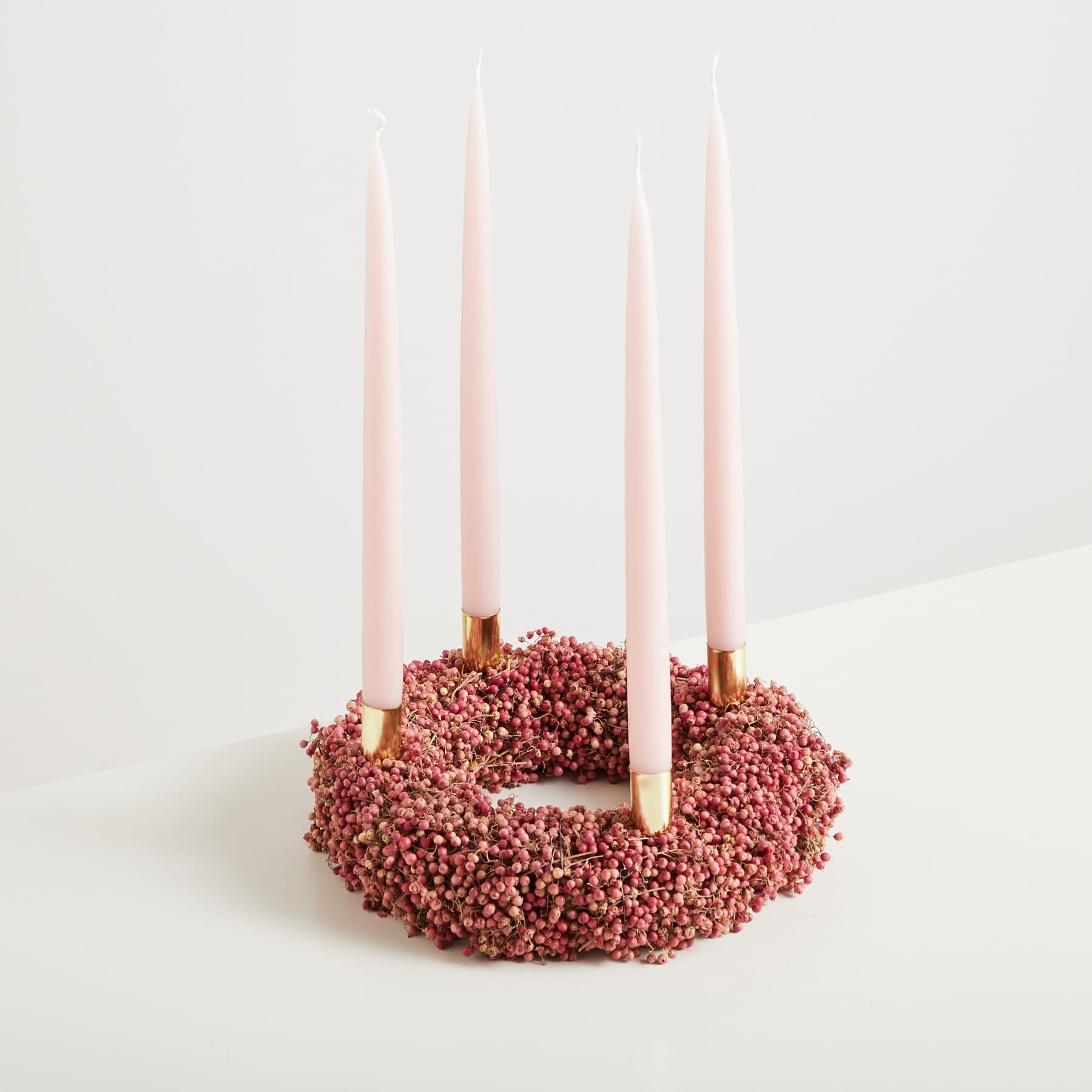 Ein kreisförmiges Arrangement aus rosa Kerzen, die auf einem Bett aus kleinen roten Beeren oder Samen vor einem schlichten weißen Hintergrund ruhen.