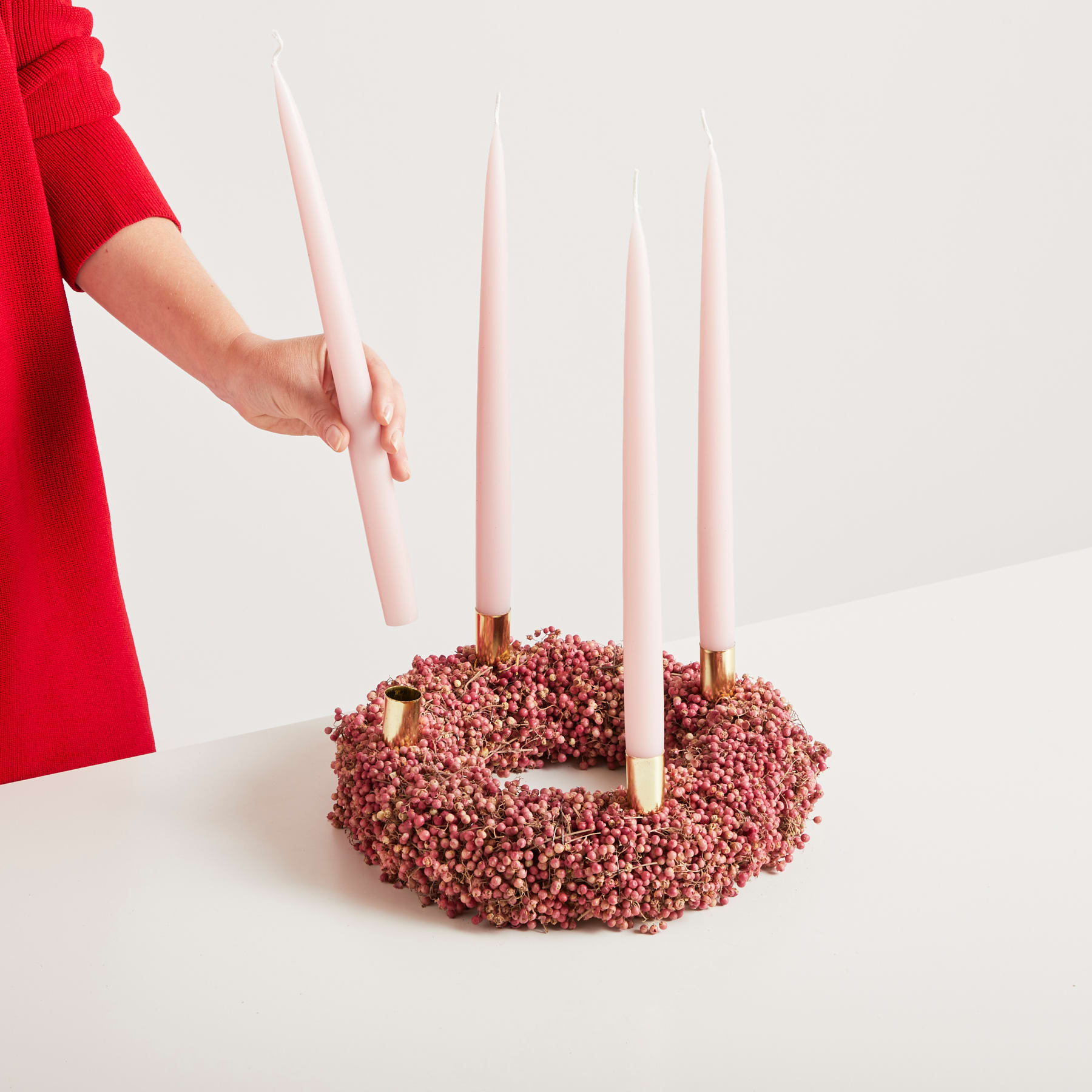 Eine rot gekleidete Hand hält rosa Kerzen auf einem kranzartigen Arrangement aus scheinbar zerdrückten rosa Blütenblättern oder Beeren vor einem schlichten weißen Hintergrund.
