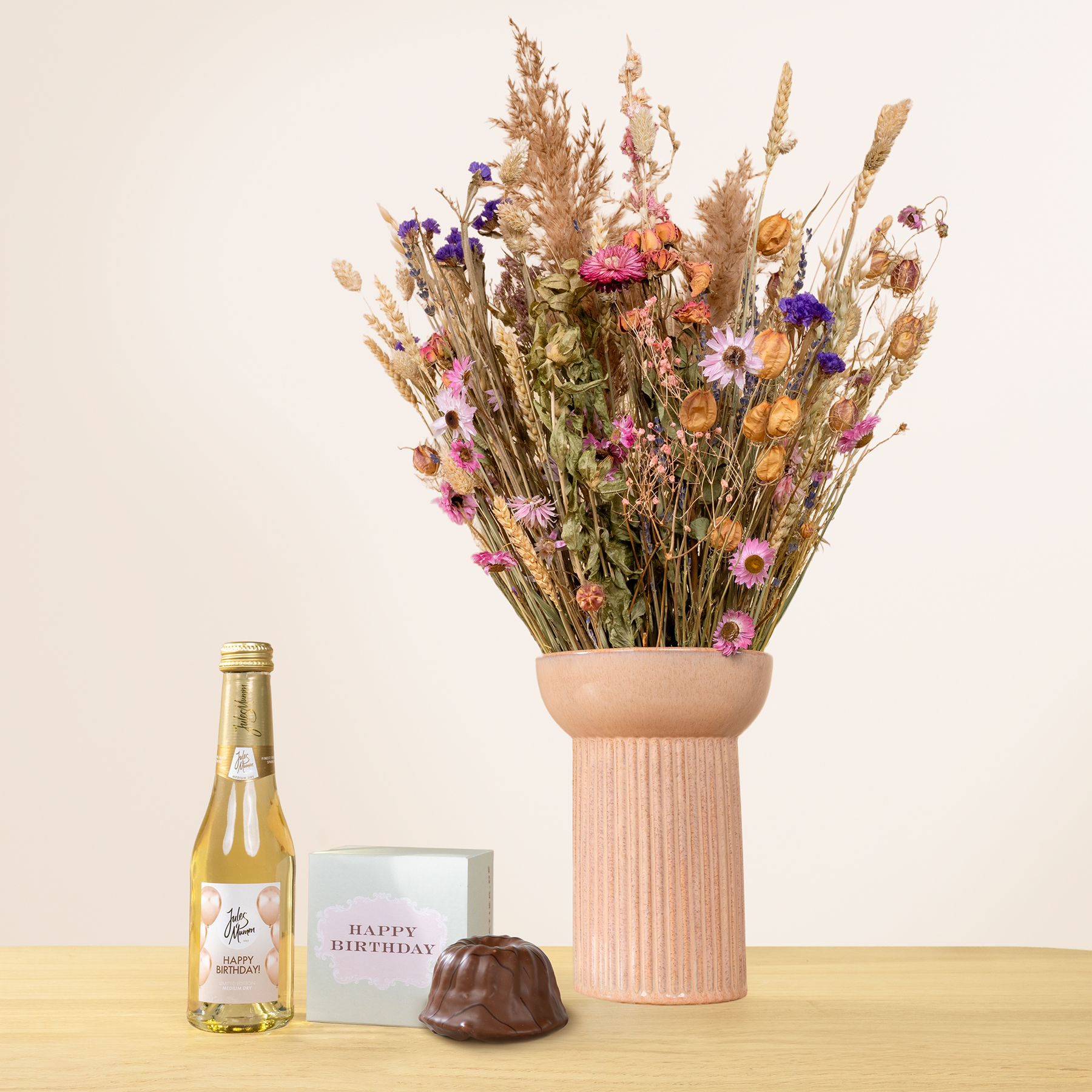 Auf einer Holzoberfläche steht eine Vase mit einem farbenfrohen Strauß getrockneter Blumen, dazu eine Flasche Wein und ein Schokoladenkonfekt.