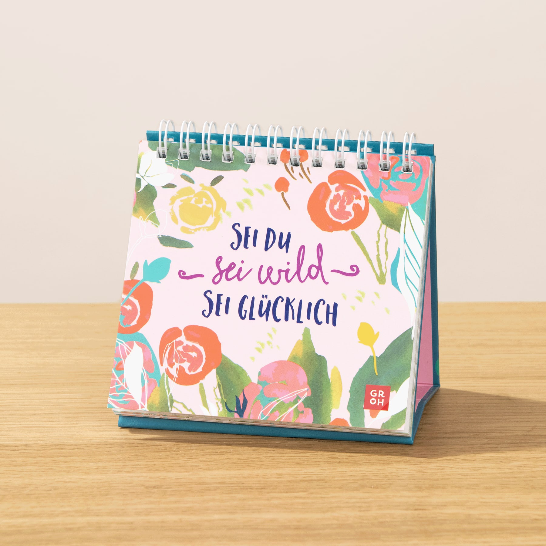 Ein farbenfroher Tischkalender mit floralen Illustrationen und dem deutschen Text „Sei du, sei wild, sei glücklich“ auf einer Holzoberfläche.