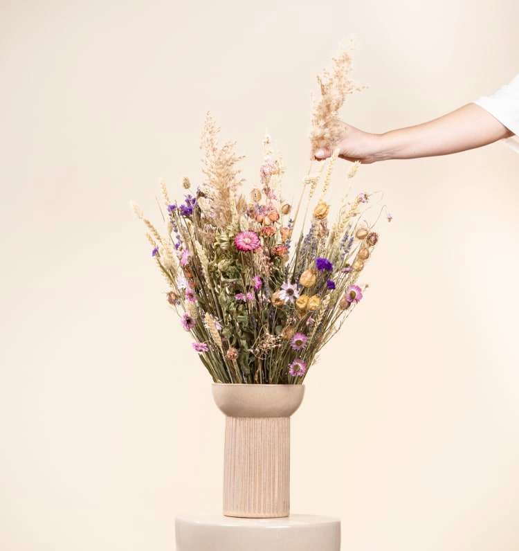Eine Hand arrangiert einen Strauß bunter Wildblumen in einer Holzvase vor einem schlichten, hellen Hintergrund.
