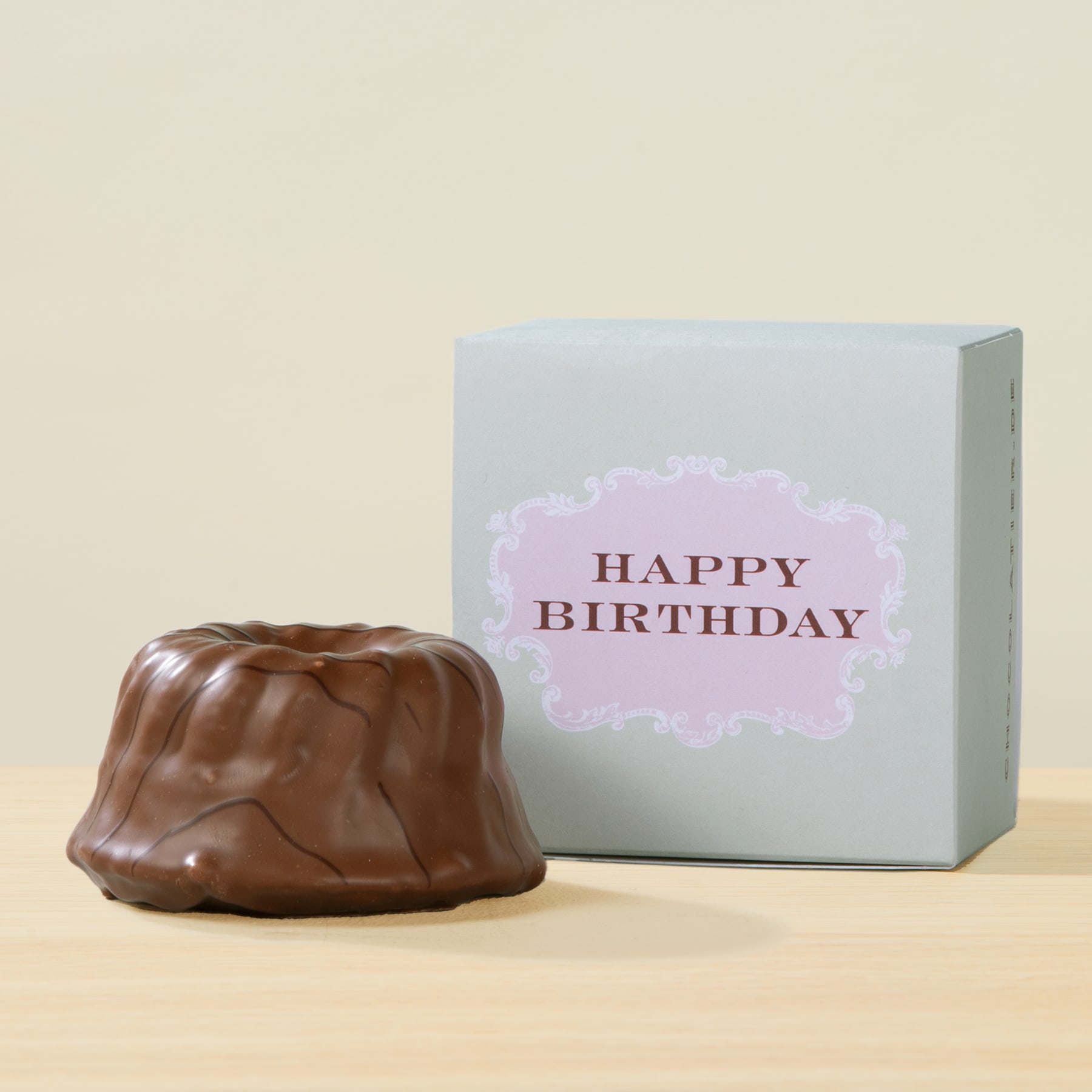 Auf einer Holzoberfläche steht ein Dessert in Form eines Schokoladenkuchens, begleitet von einer weißen Geschenkbox mit dem Aufdruck „HAPPY BIRTHDAY“.