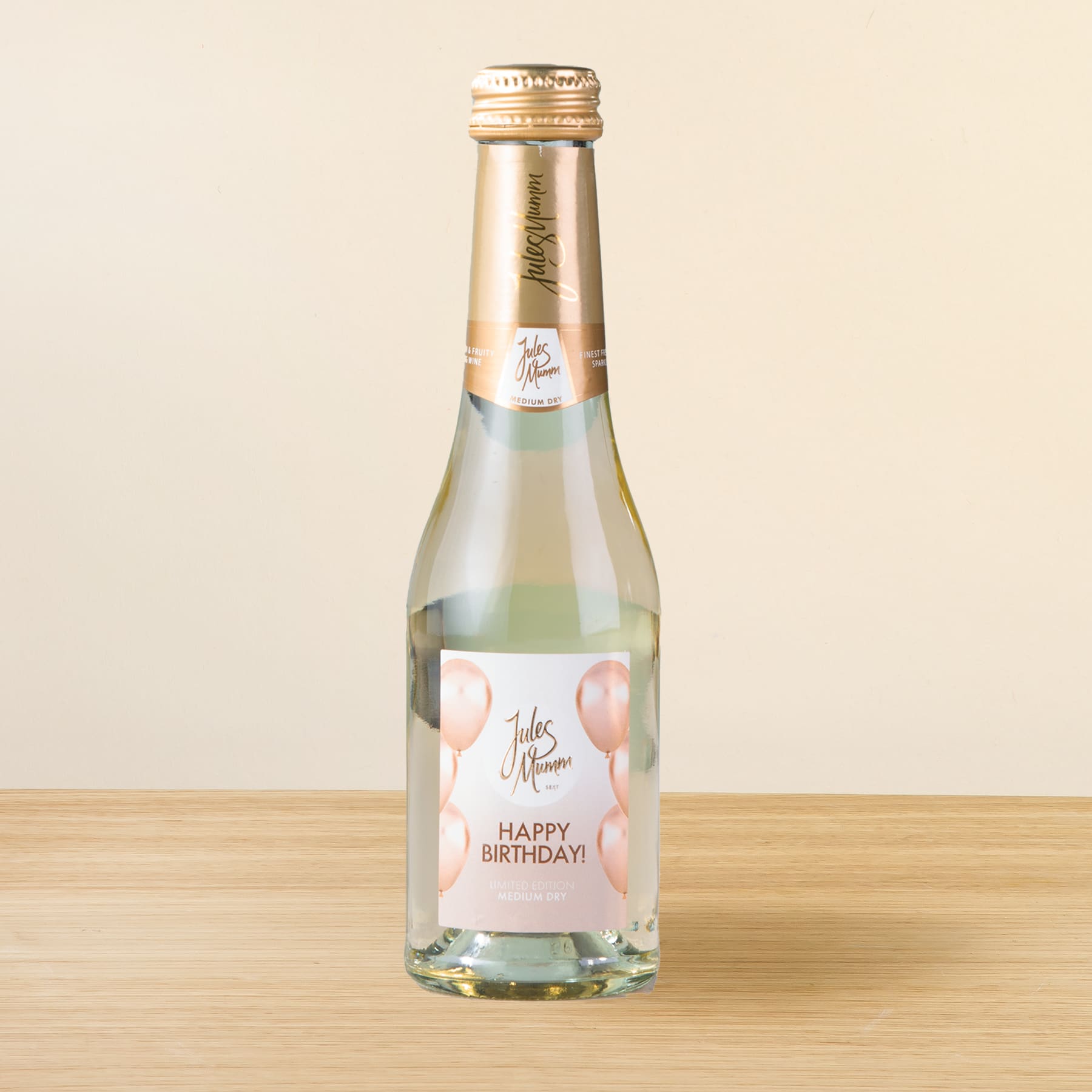 Eine Glasflasche Sekt oder Champagner mit dem Etikett „Happy Birthday!“ auf einer Holzoberfläche vor hellem Hintergrund.