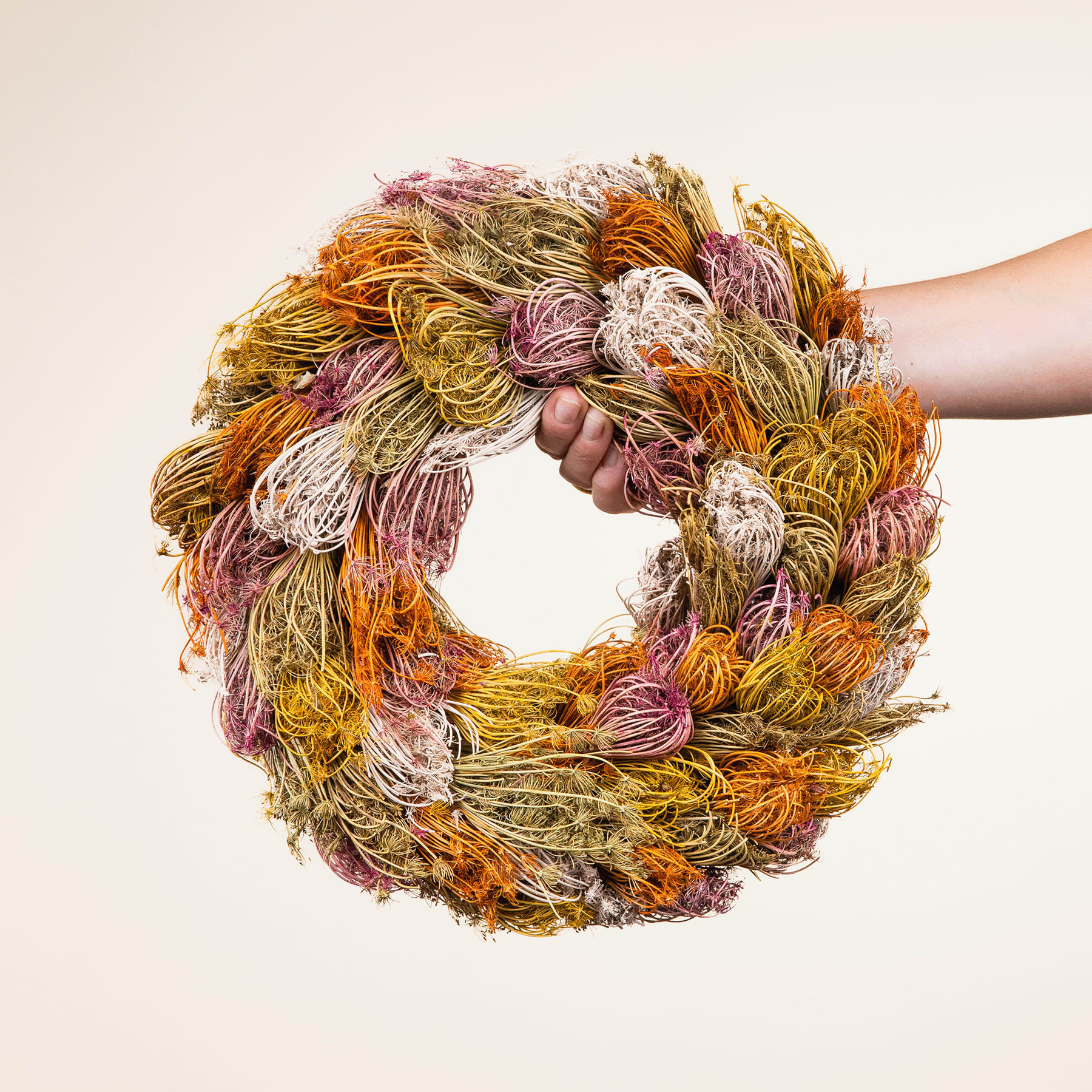 Ein farbenfroher, handgefertigter Kranz aus verschiedenen Naturfasern und Materialien in warmen Herbsttönen wird von den Händen einer Person vor einem schlichten, hellen Hintergrund gehalten.