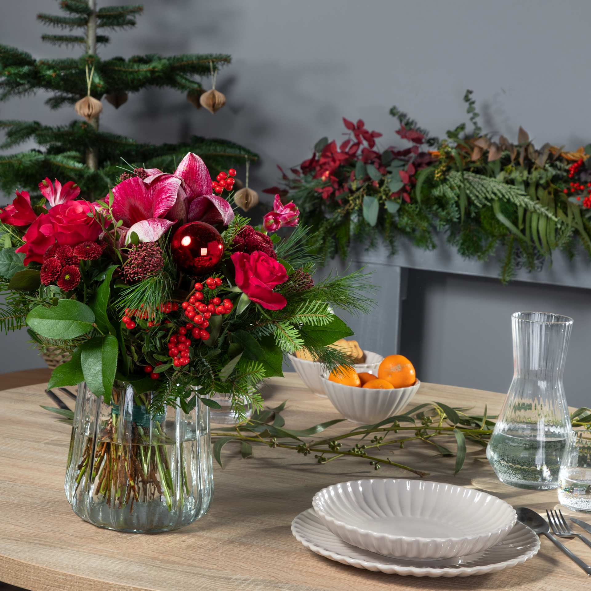 Eine festliche Tischdekoration mit farbenfrohen Blumenarrangements, Tellern und Gläsern vor dem Hintergrund eines Weihnachtsbaums und viel Grün.