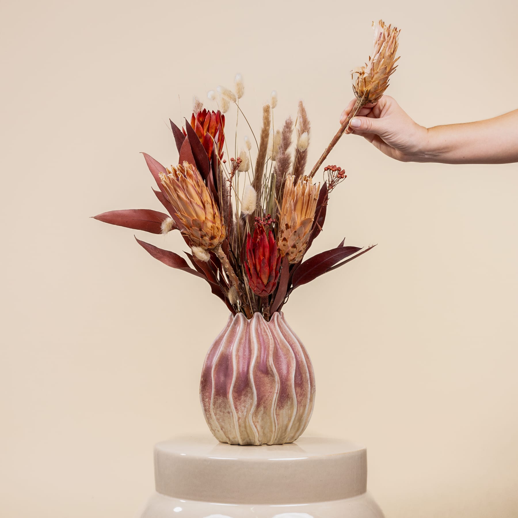 Eine Hand hält eine Vase mit einem farbenfrohen Arrangement aus getrockneten Blumen und Pflanzen vor einem neutralen beigen Hintergrund.