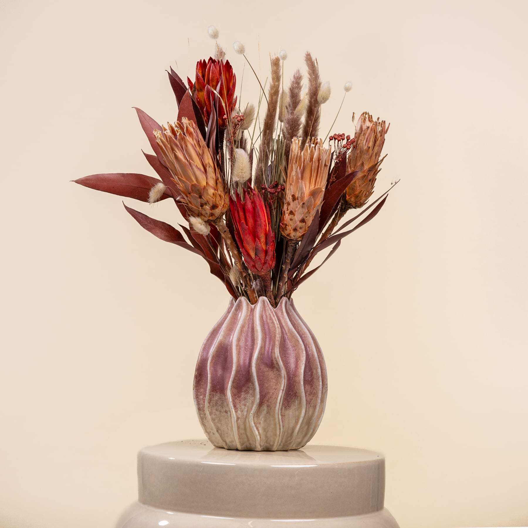 Eine Vase mit einem farbenfrohen Strauß getrockneter Blumen, darunter Proteas, Pampasgras und andere natürliche Elemente, steht auf einer schlichten, hellen Oberfläche vor einem neutralen Hintergrund.