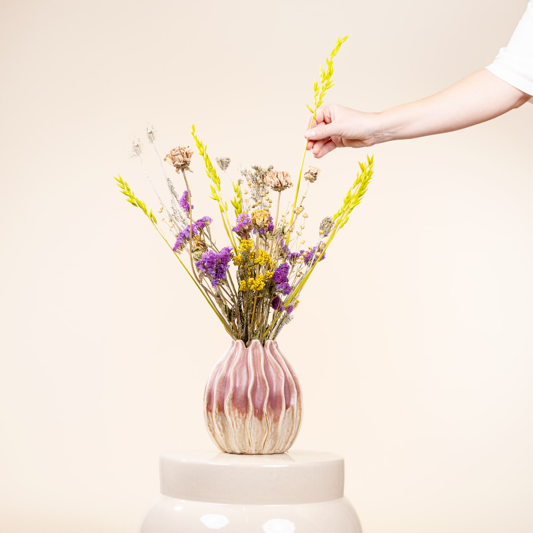 Eine Hand arrangiert einen Strauß bunter Trockenblumen in einer rosa-weißen Keramikvase vor einem schlichten beigen Hintergrund.