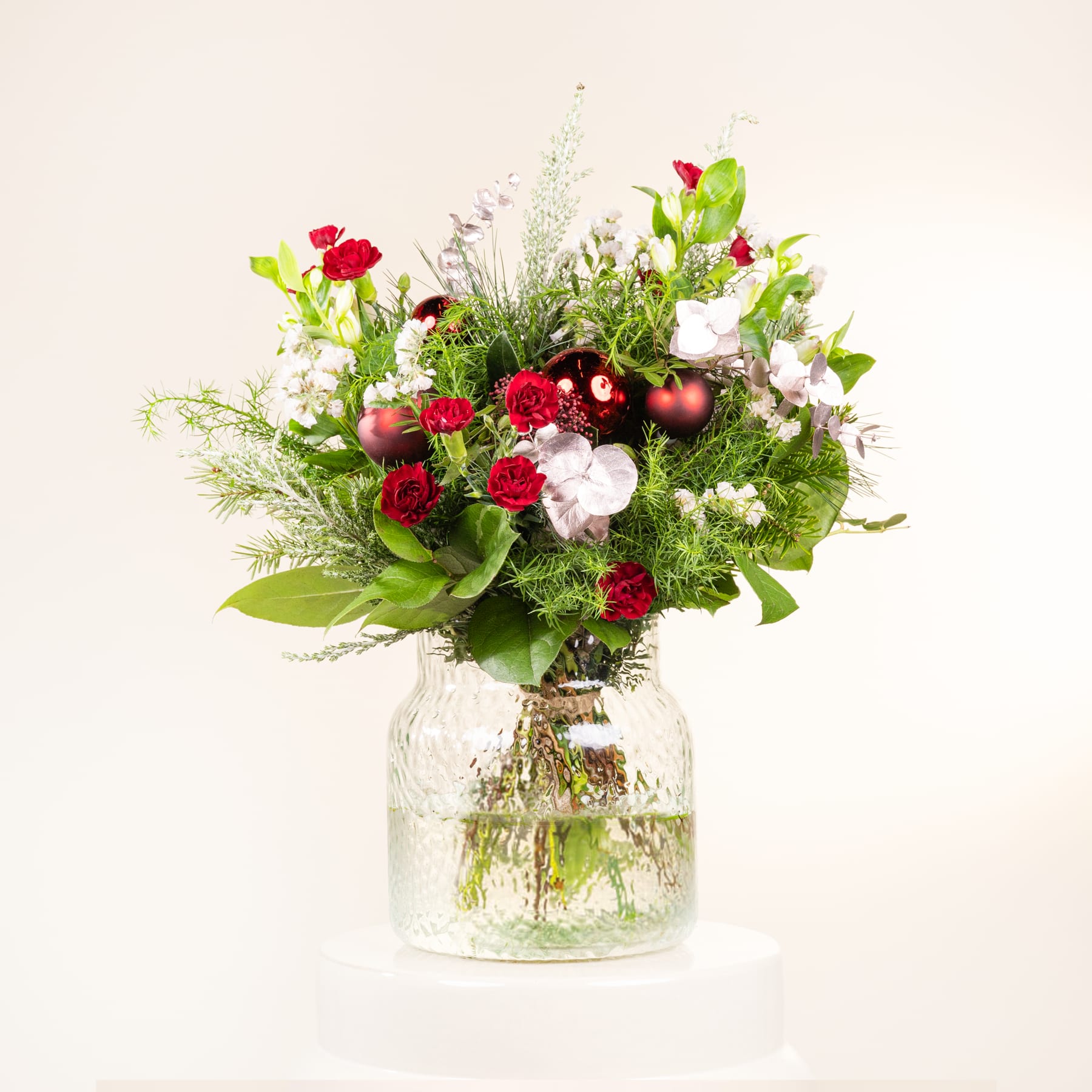 Ein leuchtender Strauß roter Rosen, weißer Nelken und Grünpflanzen, arrangiert in einem Glas vor einem schlichten Hintergrund.