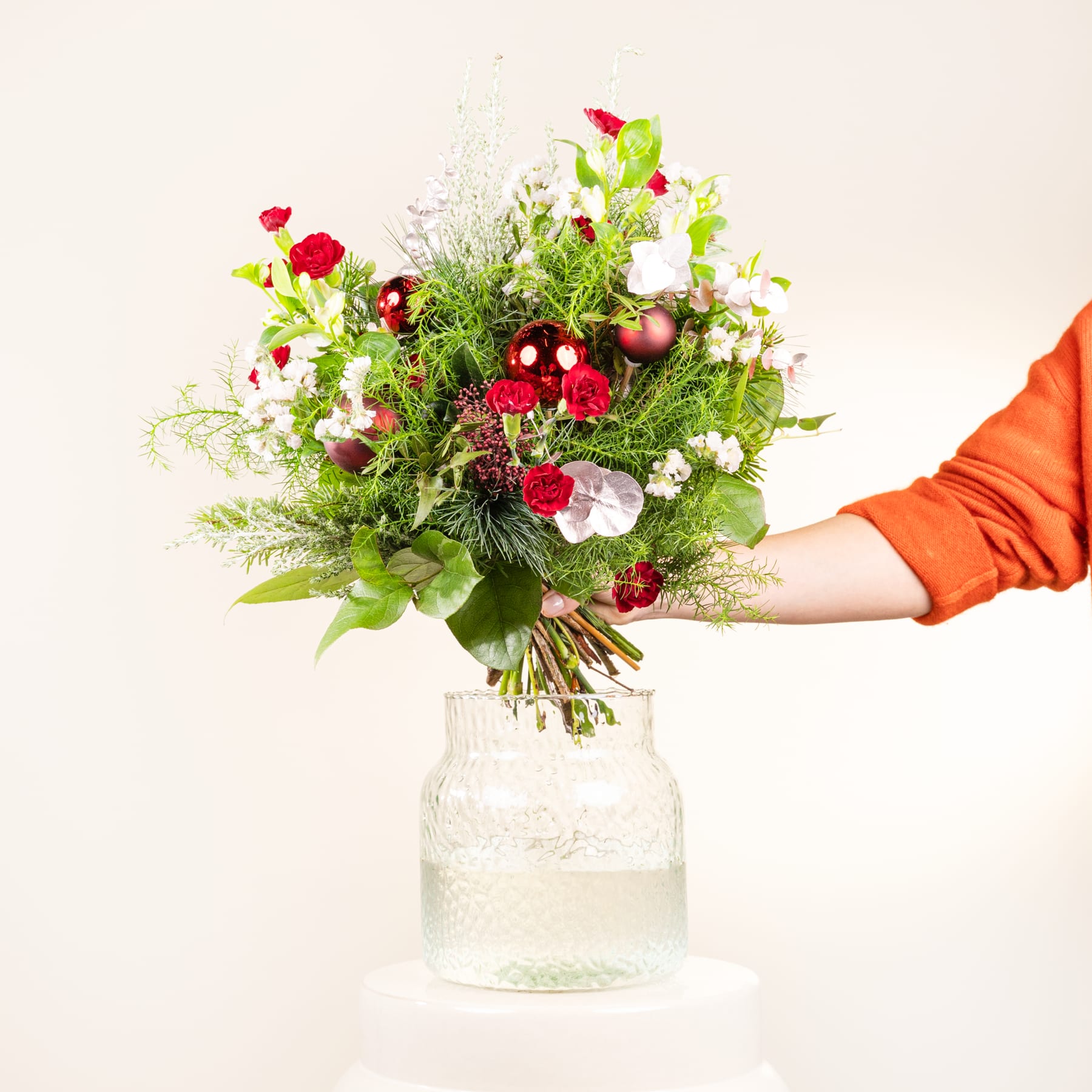 Ein farbenfroher Strauß verschiedener Blumen, darunter rote Rosen, weiße Gänseblümchen und grünes Laub, steht in einer klaren Glasvase vor einem schlichten weißen Hintergrund.
