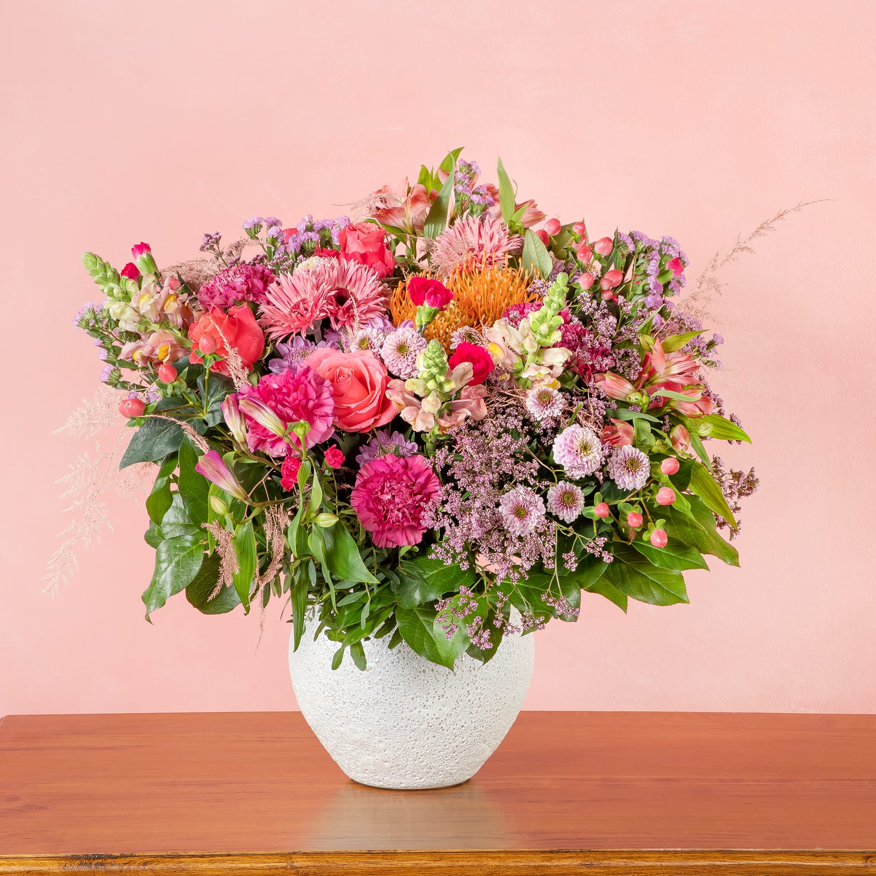 Ein lebendiger Strauß bunter Blumen, darunter Rosen, Gänseblümchen und Chrysanthemen, wird in einer weißen Keramikvase auf einem Holztisch vor einem zartrosa Hintergrund präsentiert.