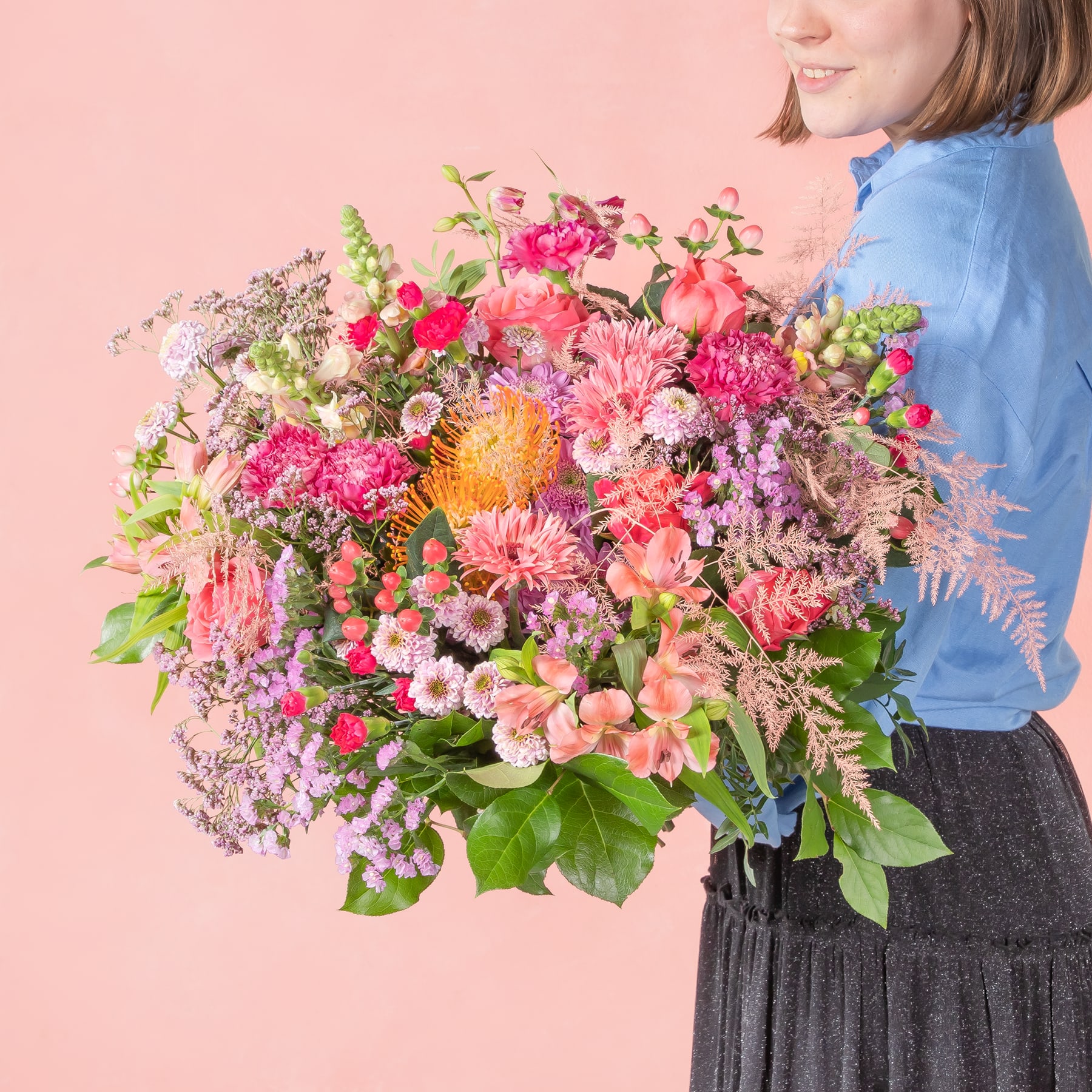 Ein leuchtender Strauß bunter Blumen, darunter rosa Nelken, gelbe Gänseblümchen und lila Flieder, wird von einer Person gehalten, die ein blaues Jeanshemd vor einem zartrosa Hintergrund trägt.