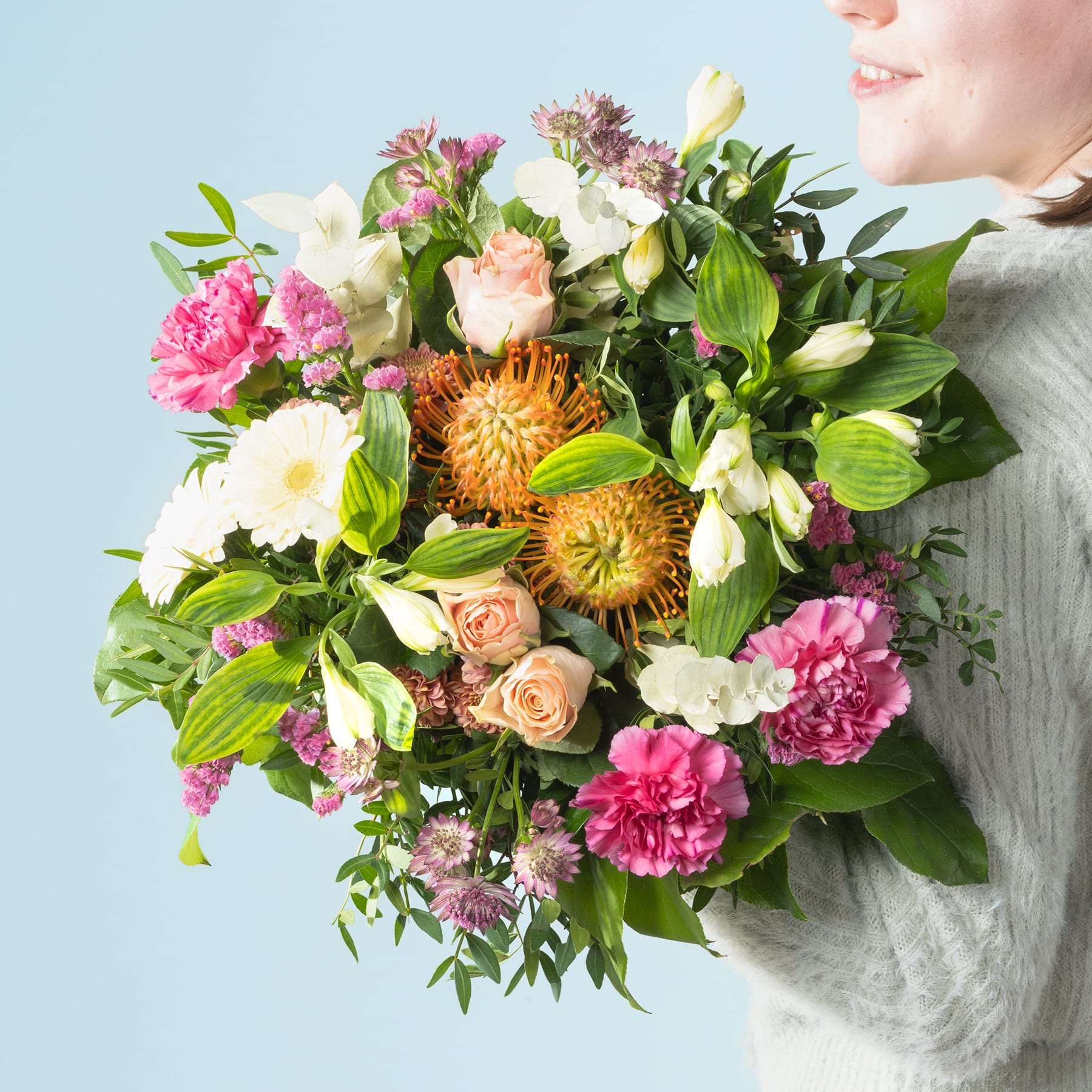 Ein farbenfroher Strauß verschiedener Blumen, darunter Pfingstrosen, Rosen und Proteas, gehalten von einer Person in einem kuscheligen Pullover vor einem sanften blauen Hintergrund.